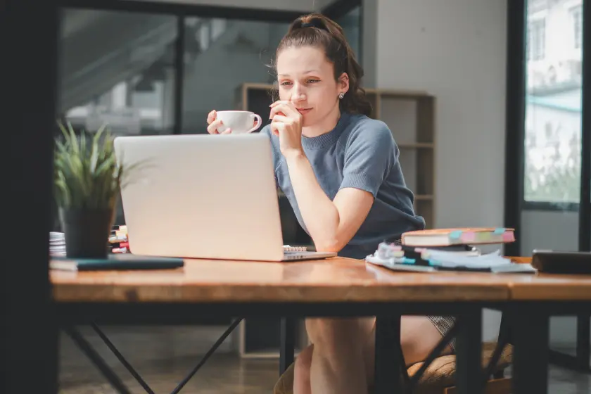 A woman is sitting at a wooden desk, looking at her laptop. She is holding a cup of coffee and appears to be focused on her work.