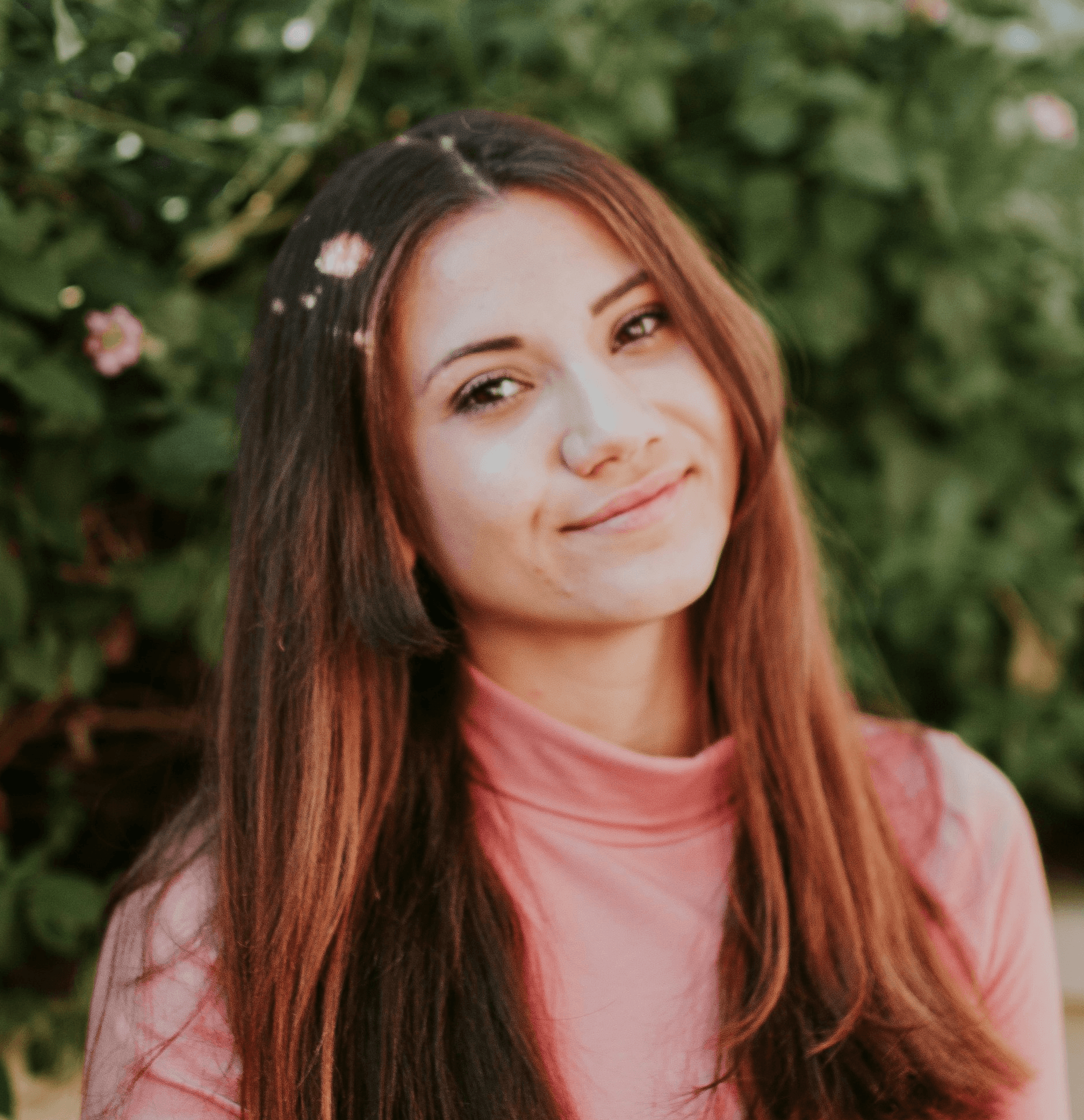 A young woman with long hair smiles softly, surrounded by greenery and delicate flowers in the background.