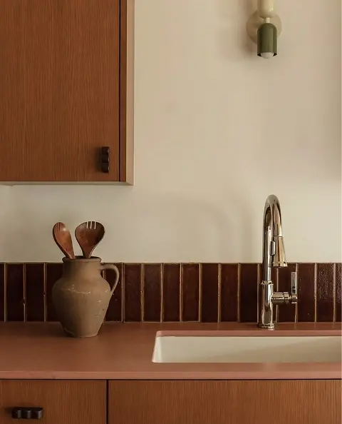 Modern kitchen corner with warm wood cabinetry, a glossy brown tiled backsplash, a stainless steel pull-down faucet over a built-in sink, and a rustic ceramic pitcher holding wooden serving spoons