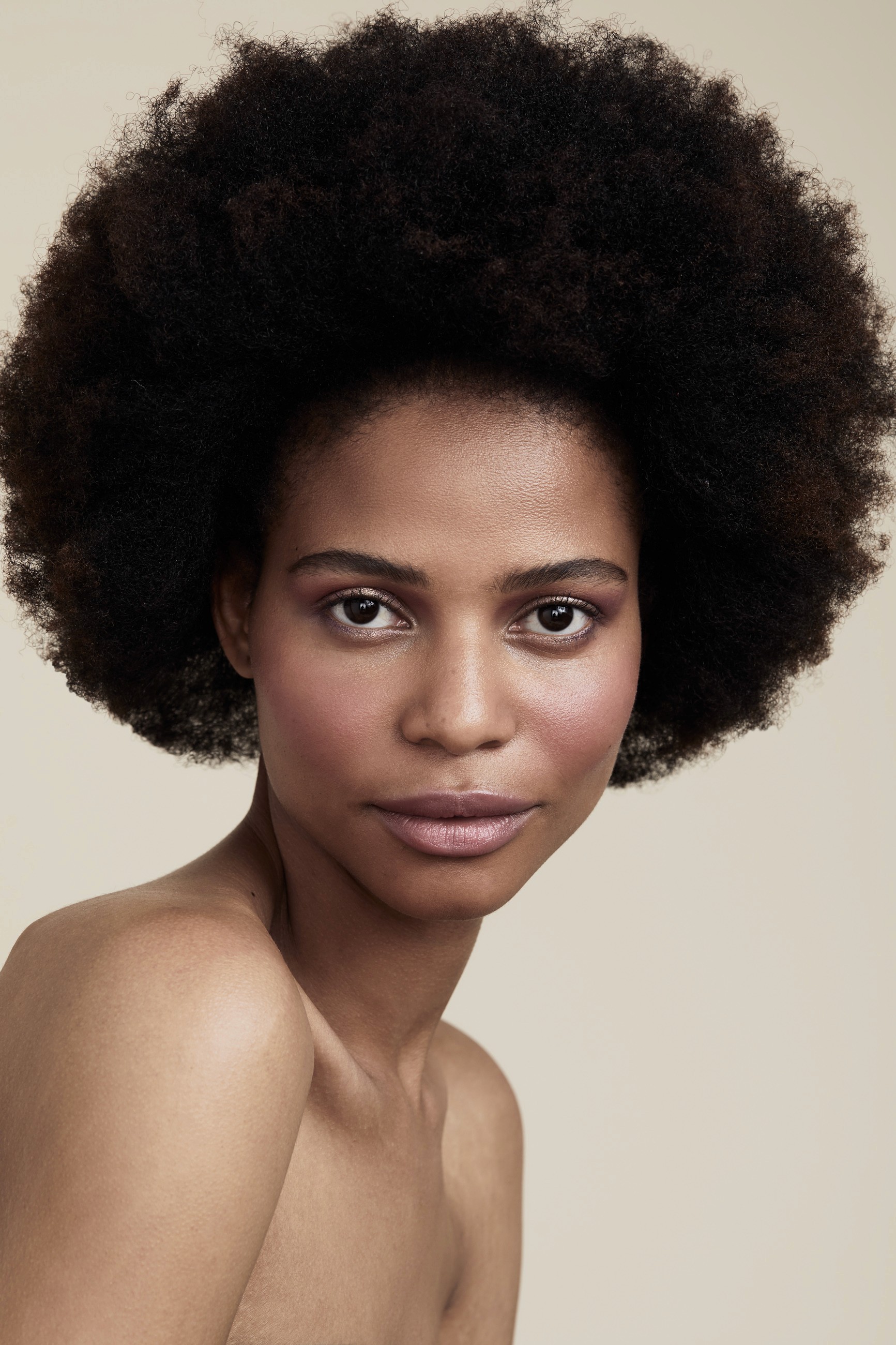 Studio beauty portrait of woman with voluminous afro and bare shoulders, featuring natural makeup and soft neutral lighting against beige backdrop.