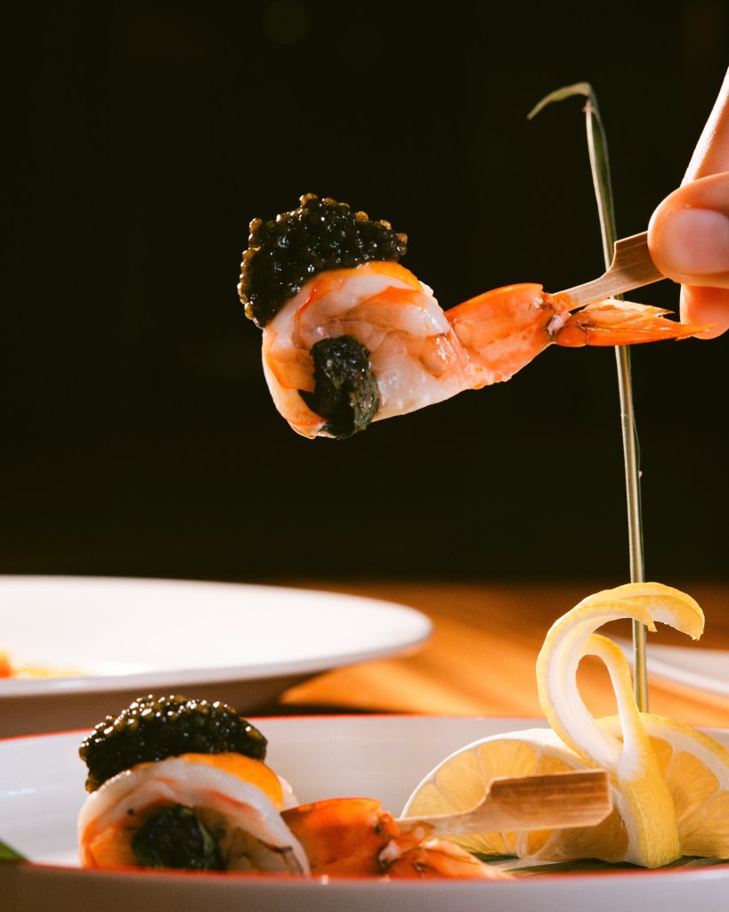 A close-up of a sushi dish featuring shrimp topped with caviar, being held by chopsticks, with slices of lemon on the side.