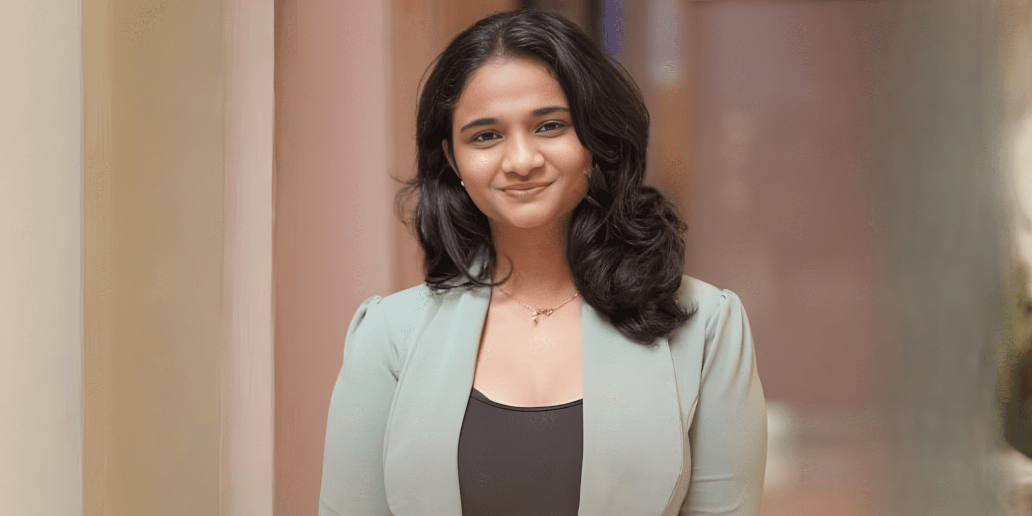 A woman with long black hair smiles warmly, wearing a light green blazer over a black top. The background is softly blurred, creating a welcoming atmosphere.