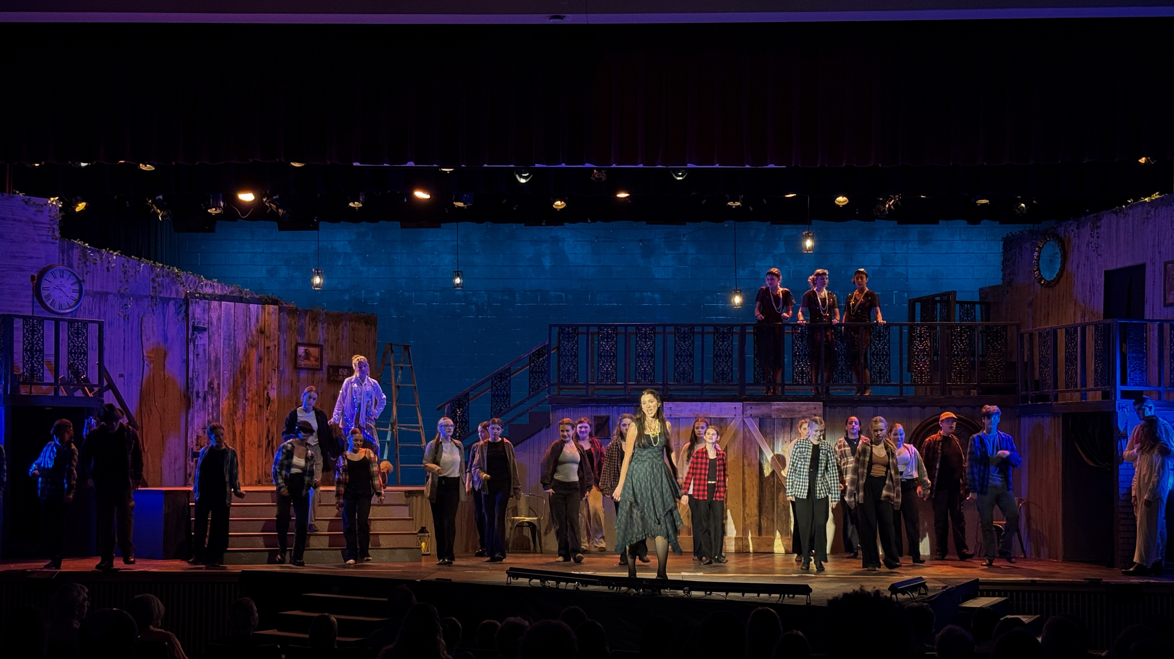 A theatrical production captures attention with amber top lighting and a blue backdrop, as Persephone stands prominently on the stage thrust, flanked by a diverse cast ensemble.