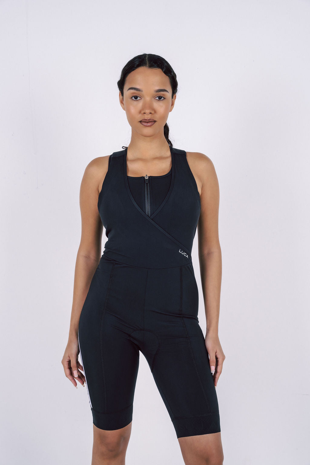 A-studio-shot-of-a-woman-modeling-a-black-women's-cycling- all-in-one-against-a-plain-white- background-The-suit-is-sleeveless- and-features-a-racerback-design- with-a-black-inner-layer-visible- below-the-shoulders-The-front- includes-a-short-zipper-and-a- defining-crossover-V-neck-detail- The-integrated-shorts-have-a-mid-thigh-length-and-a-thin-white-stripe-detail-on-the-lower-outer-side-of-the-lef- leg