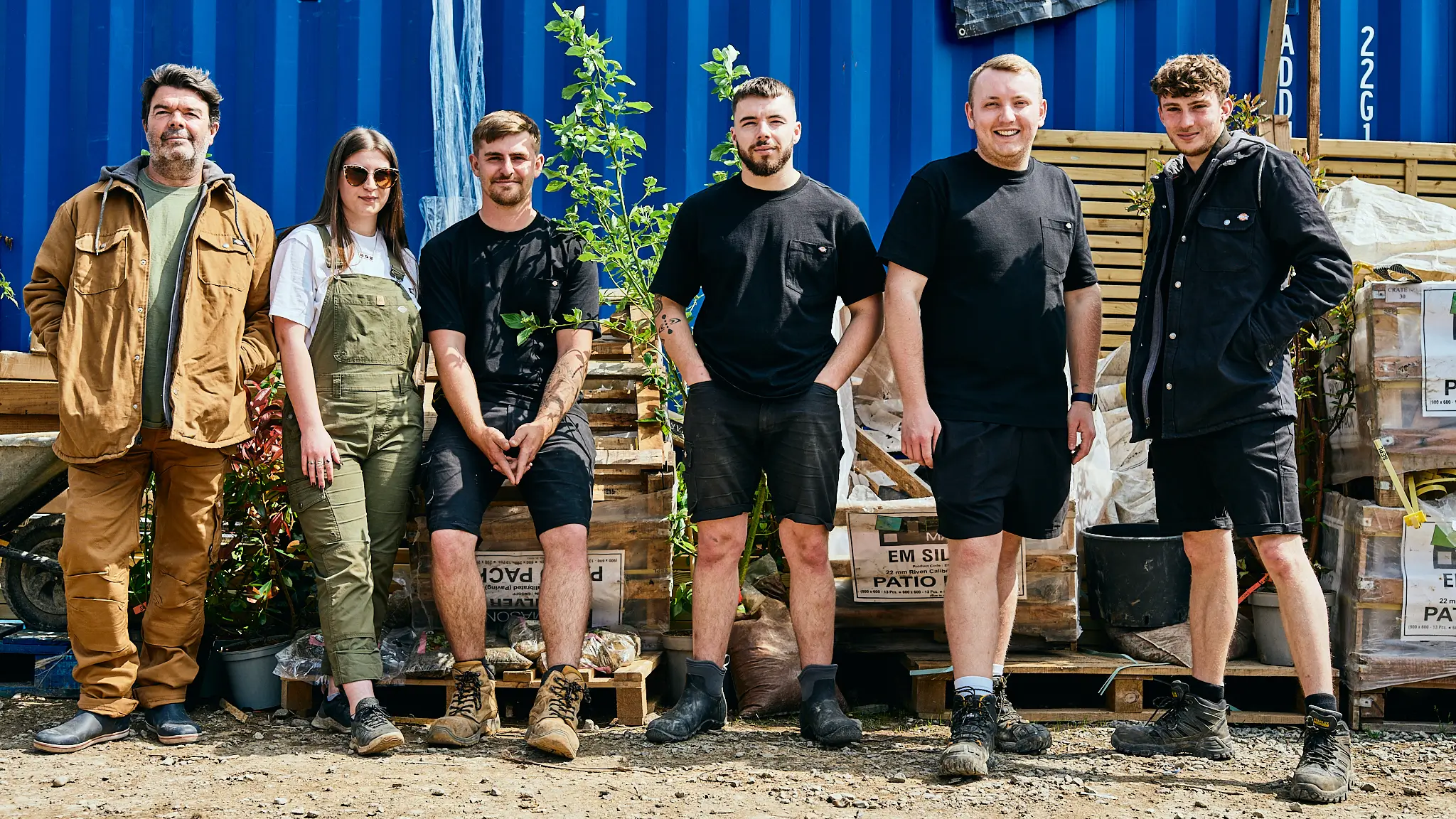 Five individuals stand together outdoors, wearing a mix of military-style and casual clothing, with trees behind them.