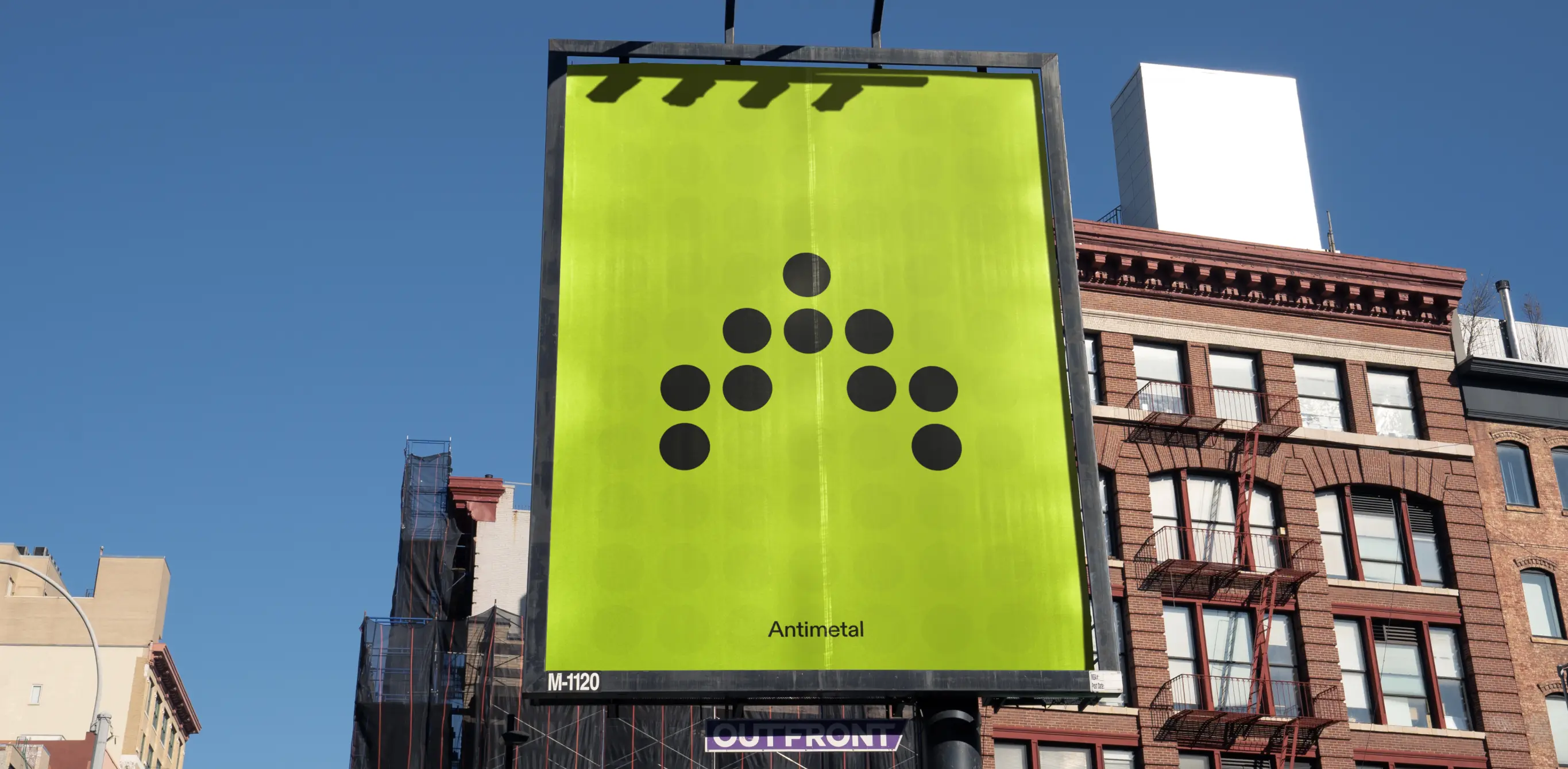 A bright green billboard with a minimalist design featuring black dots arranged in a pattern and the word 'Antimetal' at the bottom. The billboard is mounted on a structure in an urban area, with buildings and a clear blue sky in the background.