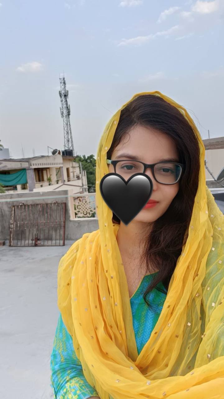 Smiling young woman with long hair standing against a dark green background, holding a finger to her chin.