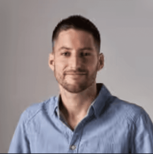 A smiling person with long, curly hair stands with arms crossed, in a modern, blurred background.