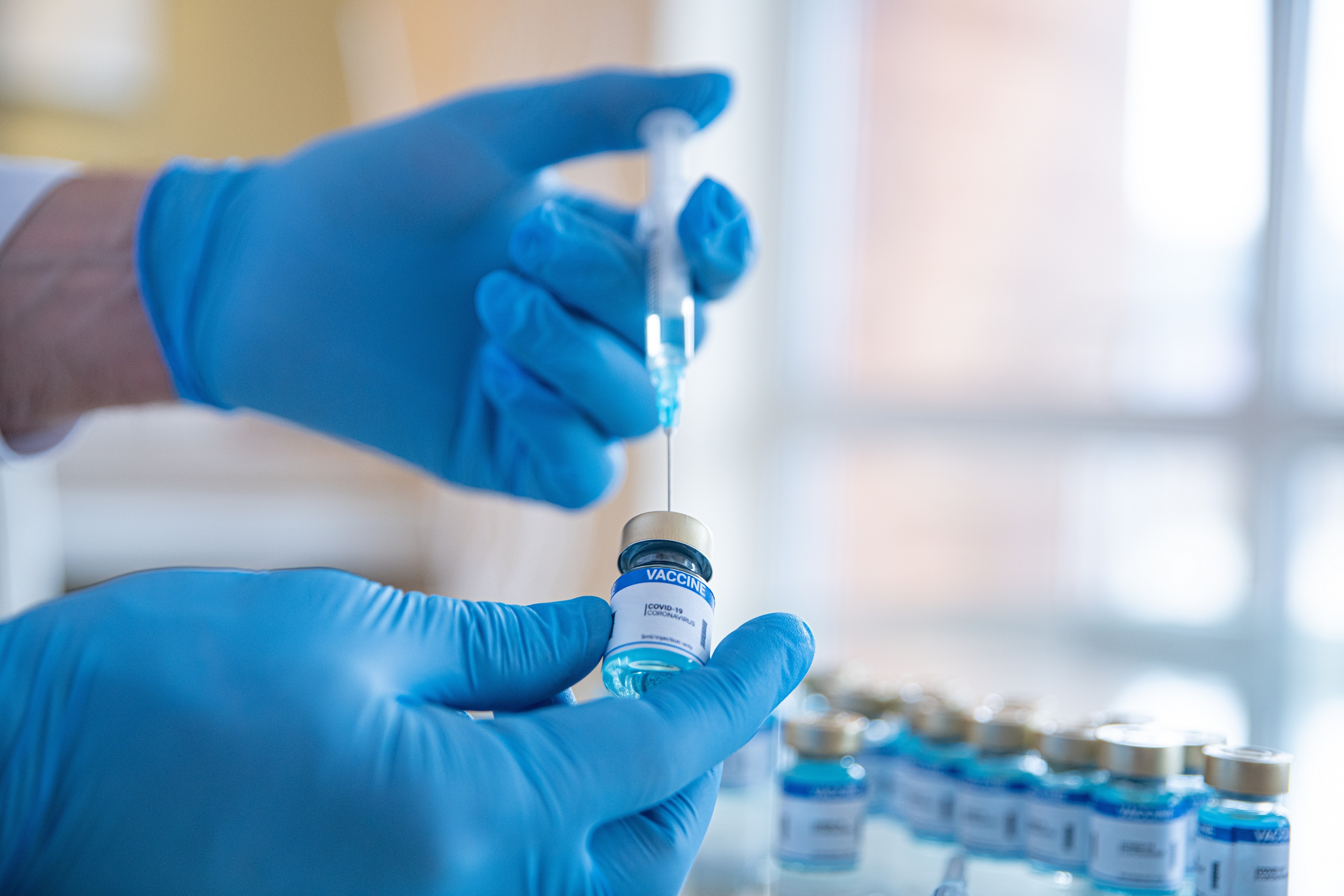 Hands in blue gloves extracting a vaccine liquid out of a vial labelled "vaccine" using a syringe.