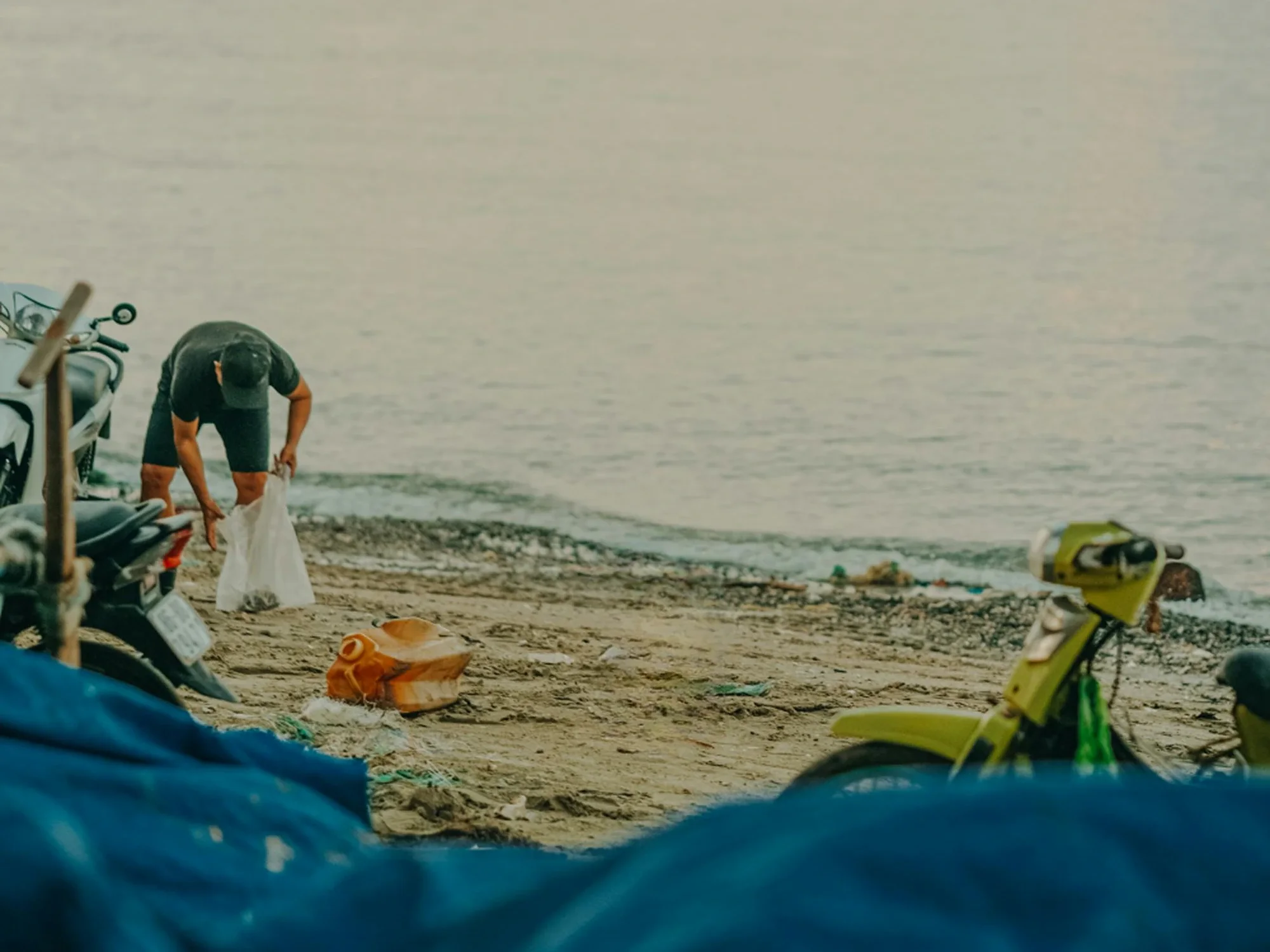 A person wearing a cap bends over, picking up trash in a white bag on a sandy beach.