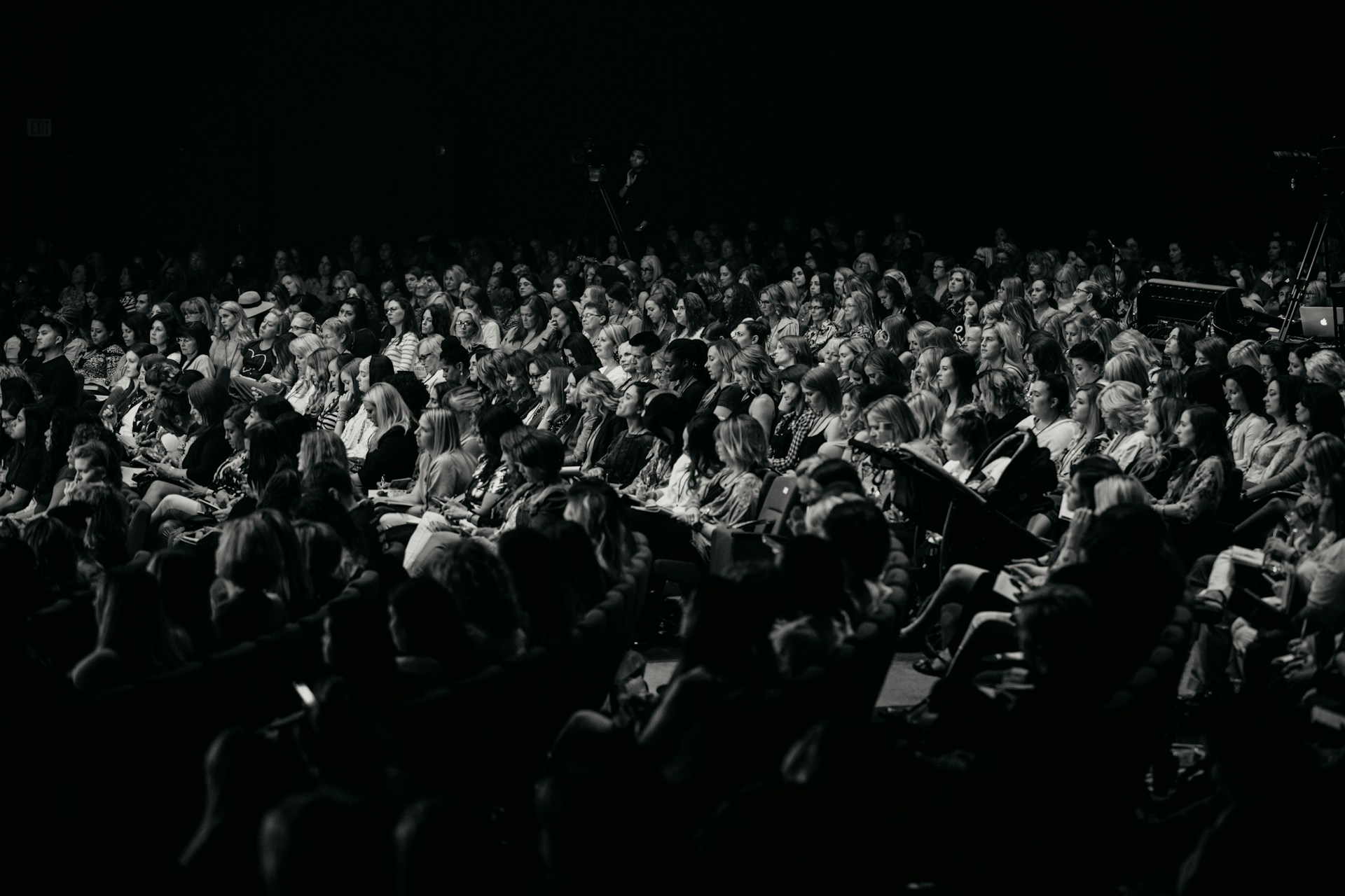 A wide, low-angle shot of a dark auditorium filled with an audience watching a live performance on a large, glowing stage. The high-contrast, atmospheric lighting highlights the scale of the event, serving as a visual payoff for the "8:00 AM Stress Test"—the moment a complex design system successfully translates from the studio to a high-density, live environment.