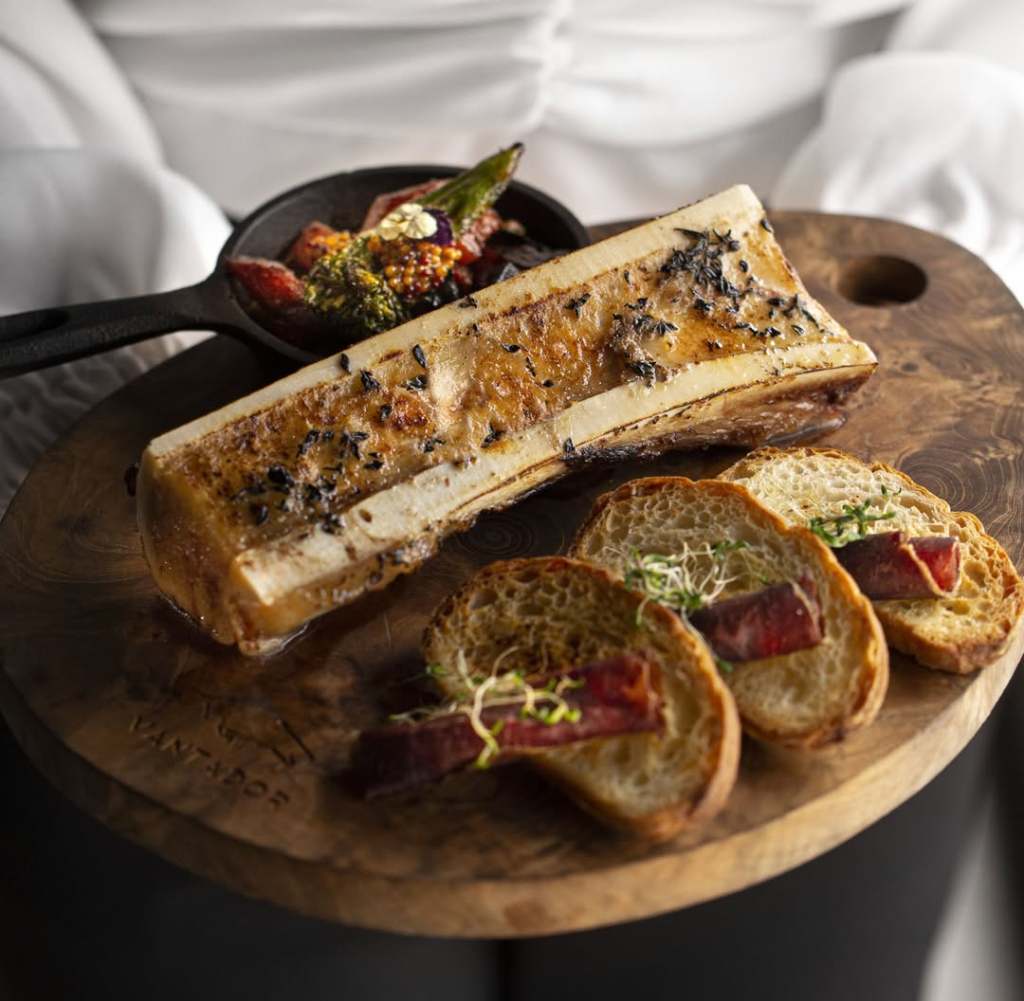 A wooden board featuring a roasted bone marrow dish, accompanied by slices of bread topped with cured meat and sprouts, with a small cast iron skillet holding colorful vegetables.