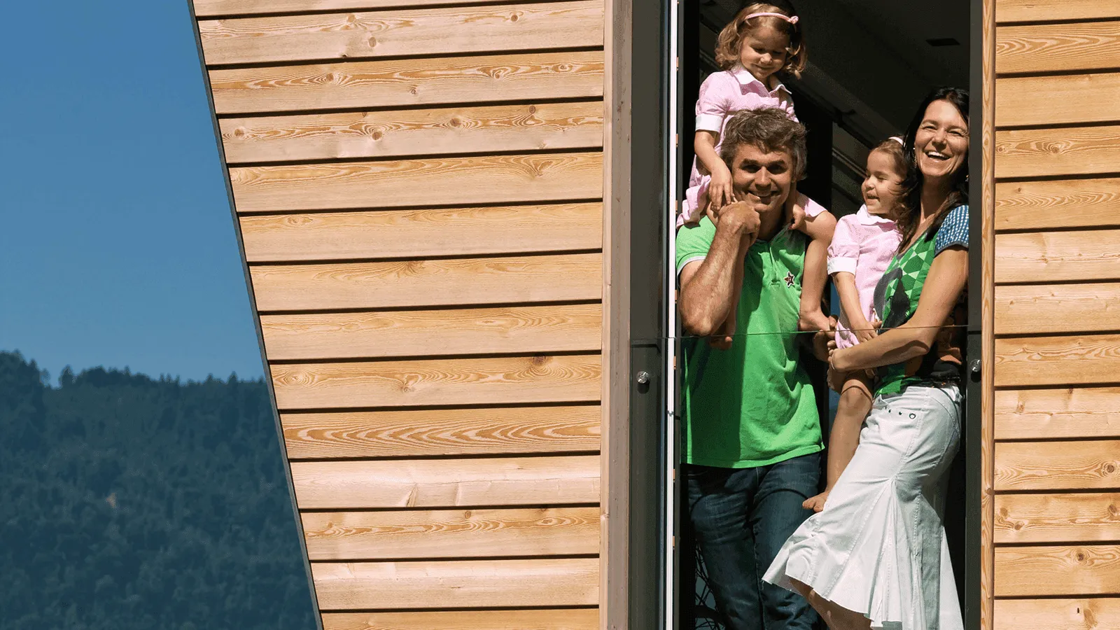 Fröhliche Familie mit zwei Kindern steht im Türrahmen eines Holzhauses mit Bergblick im Hintergrund.
