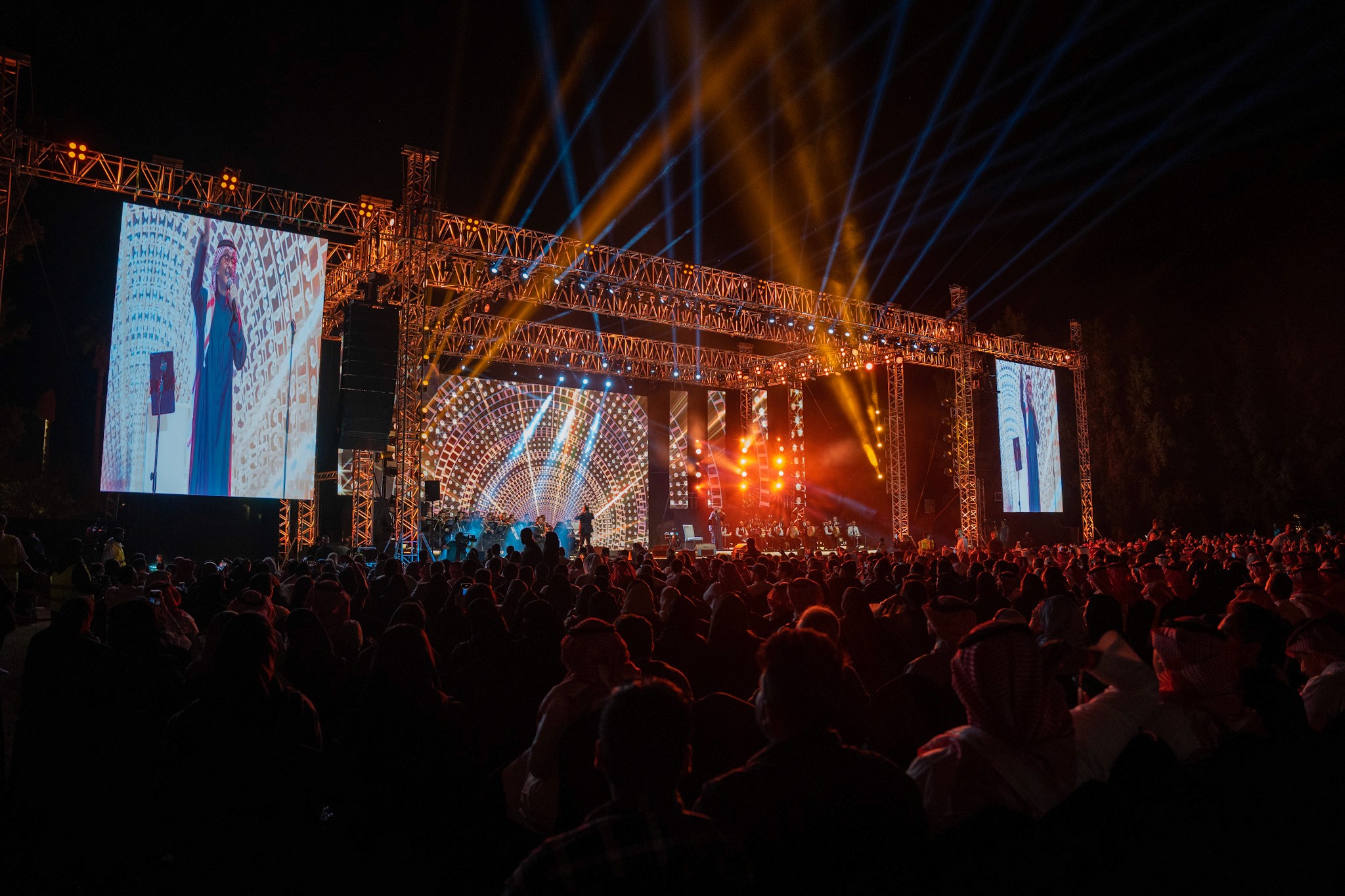 Large outdoor concert stage with illuminated screens and beams of blue and amber light above the audience