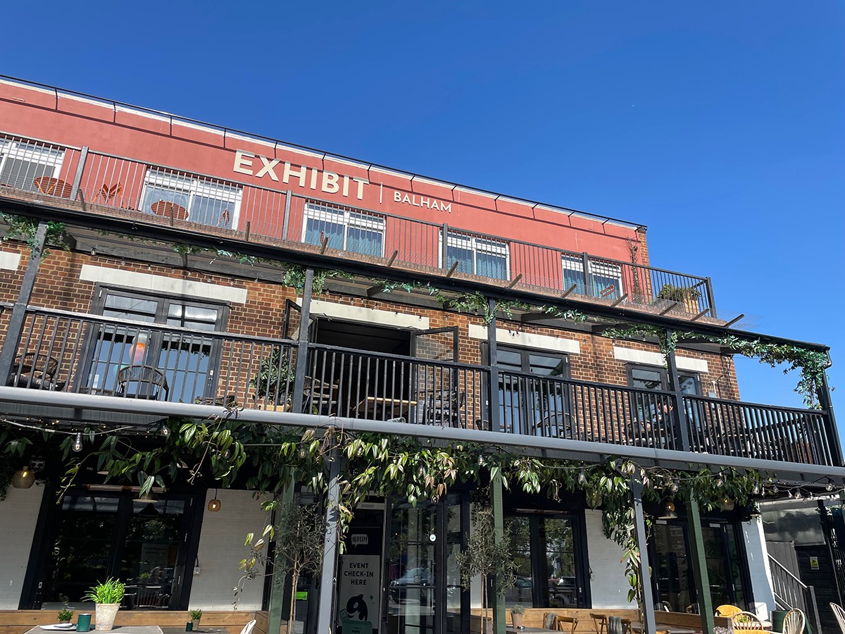 Exterior of Exhibit Balham event venue on a sunny day, featuring a red brick building with balconies, greenery and an event check-in sign at the entrance