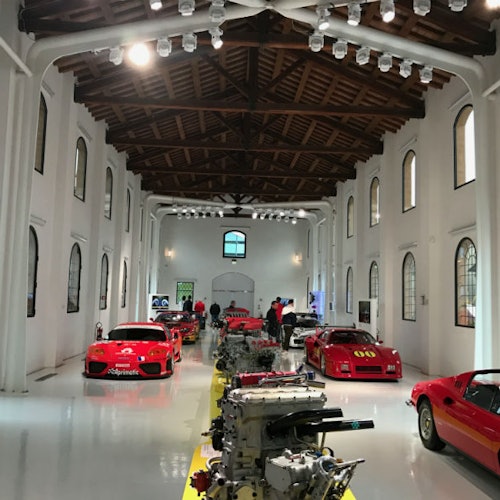 תצוגה של מכוניות אדומות ומנועים בתוך אולם מואר היטב עם קירות לבנים, חלונות קשתיים ותקרה מעץ. אנשים צופים במוצגים.