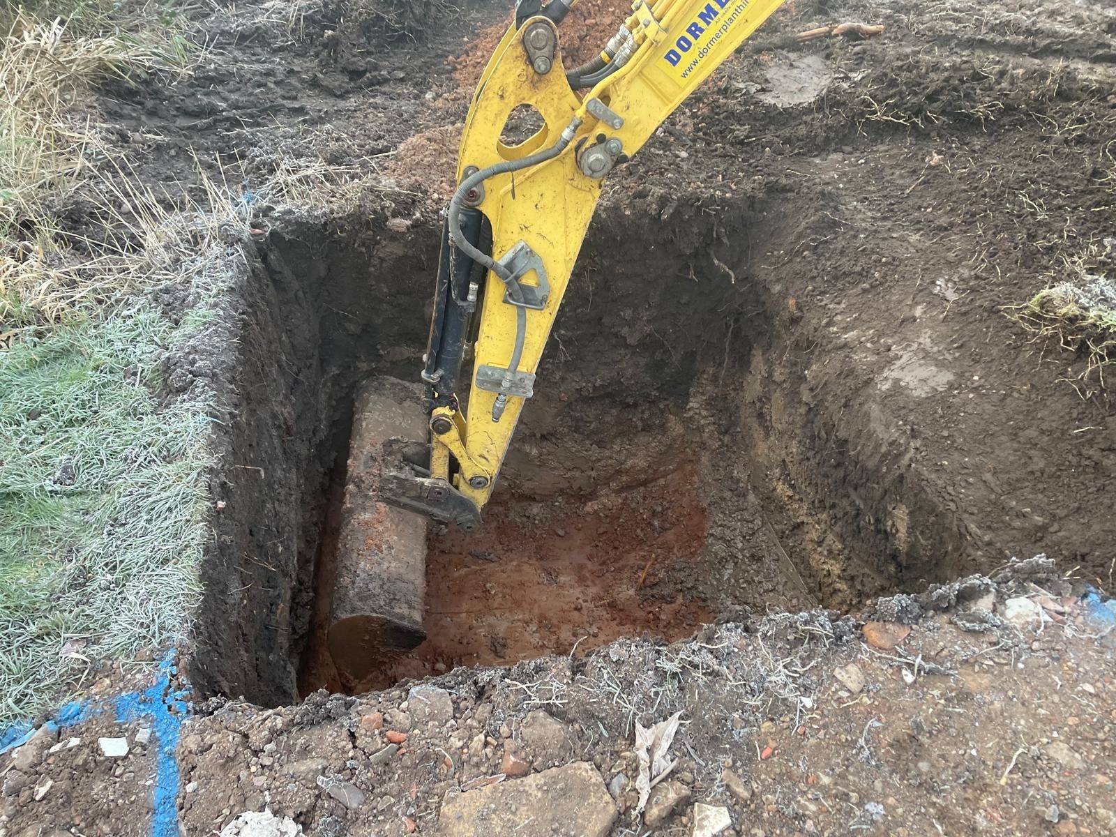 A yellow excavator is digging into a deep trench, revealing dirt and debris around the edges.