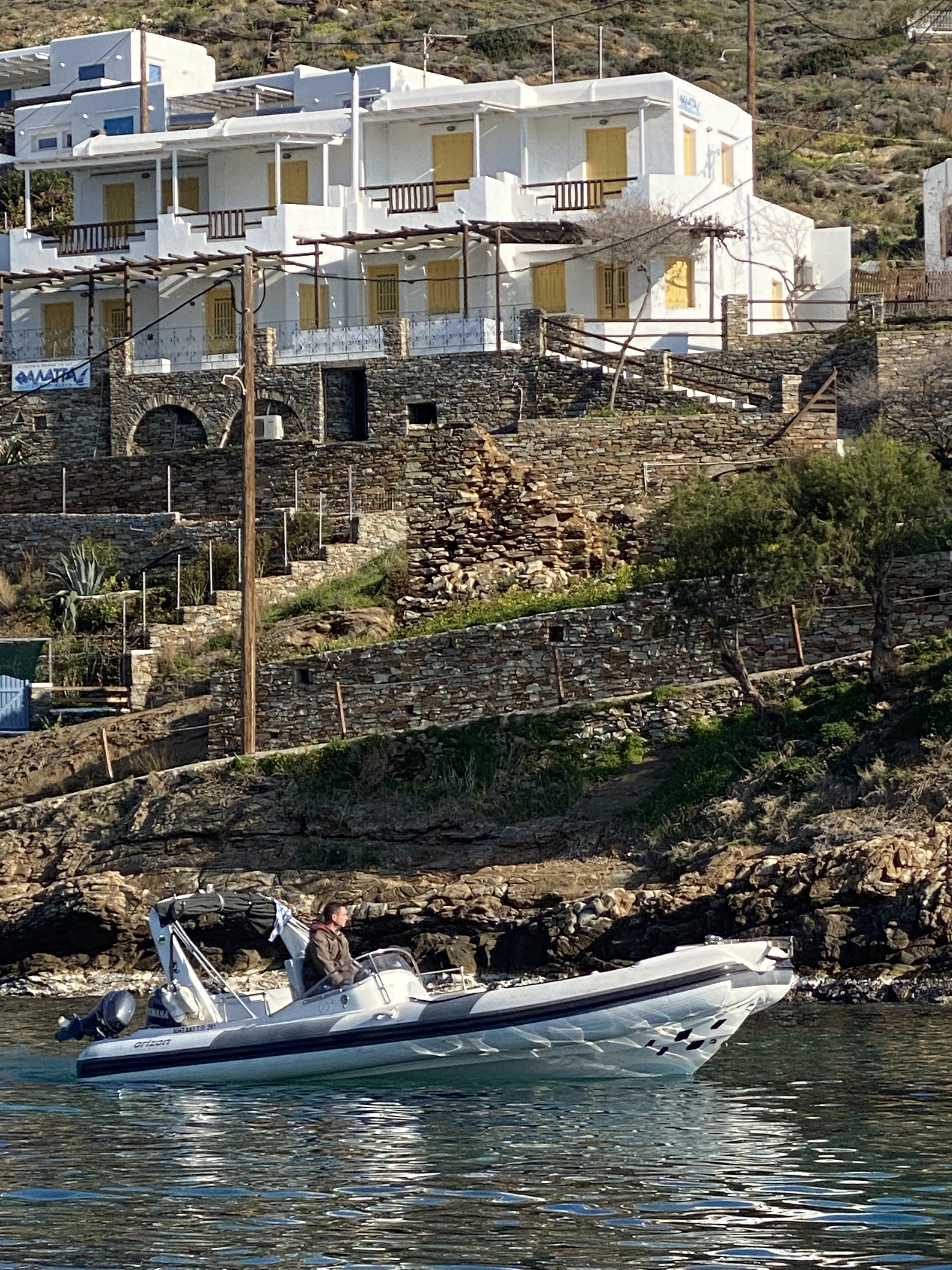 White speedboat with outboard motor cruising in calm waters near traditional Cycladic stone buildings and terraced hillside.