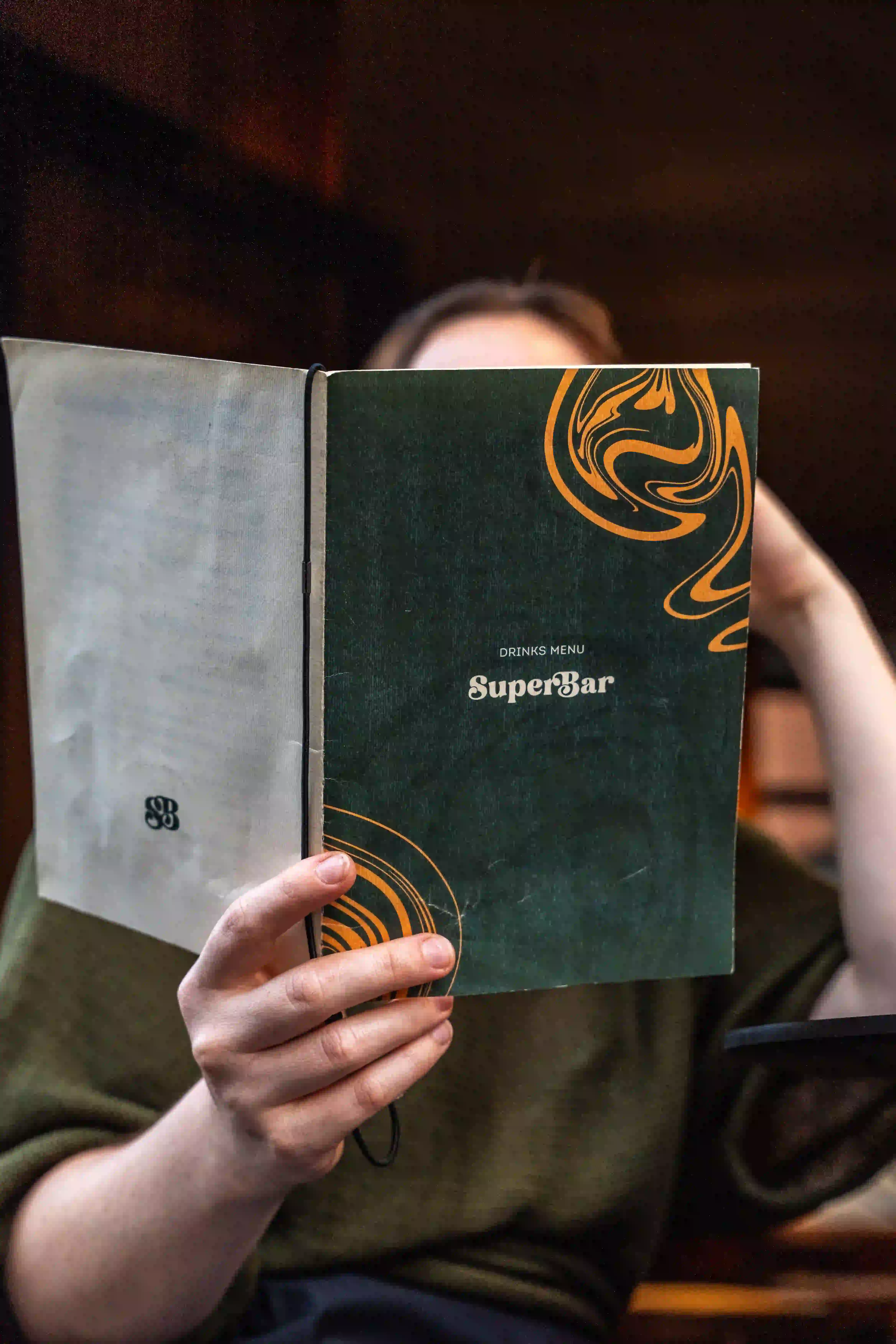 A person sits in a dimly lit restaurant, holding up an elegantly designed menu with "Super Bar" written on the front, featuring green and orange swirling patterns.
