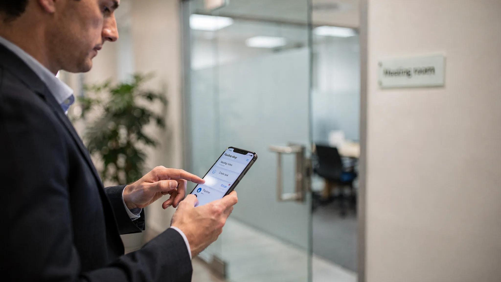 Üzleti helyzet: értékesítő egy ügyféltalálkozó után a telefonján rögzíti a meeting jegyzetét, feladatot hoz létre és frissíti a pipeline státuszt; a háttérben tárgyalóterem ajtaja, letisztult, professzionális környezet.