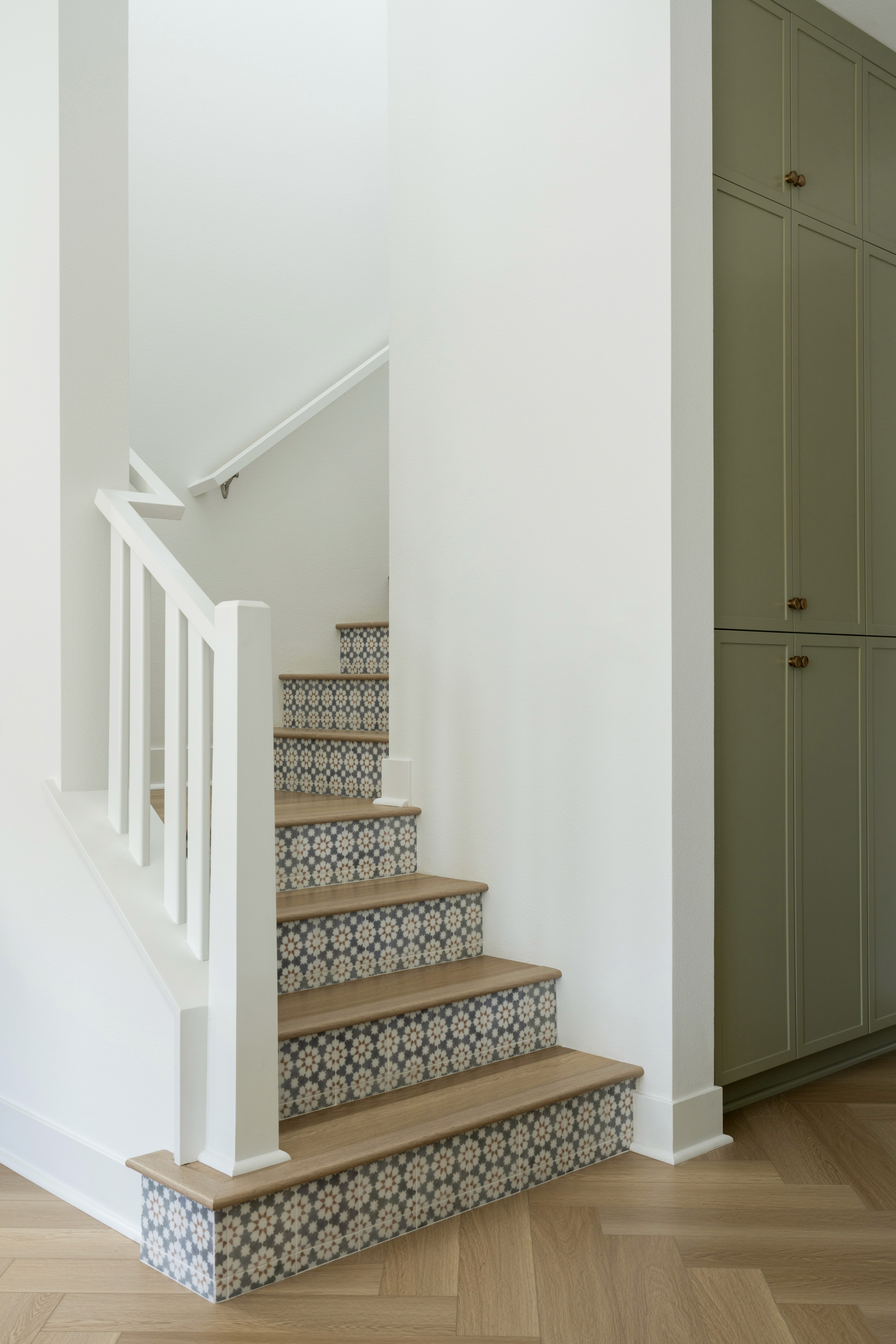 Walkway and staircase with white walls, sage green floor to ceiling cabinetry, white banister, and black and white patterned risers and wood tread matching chevron wood floors by Jubilee Interiors in Arcadia, California