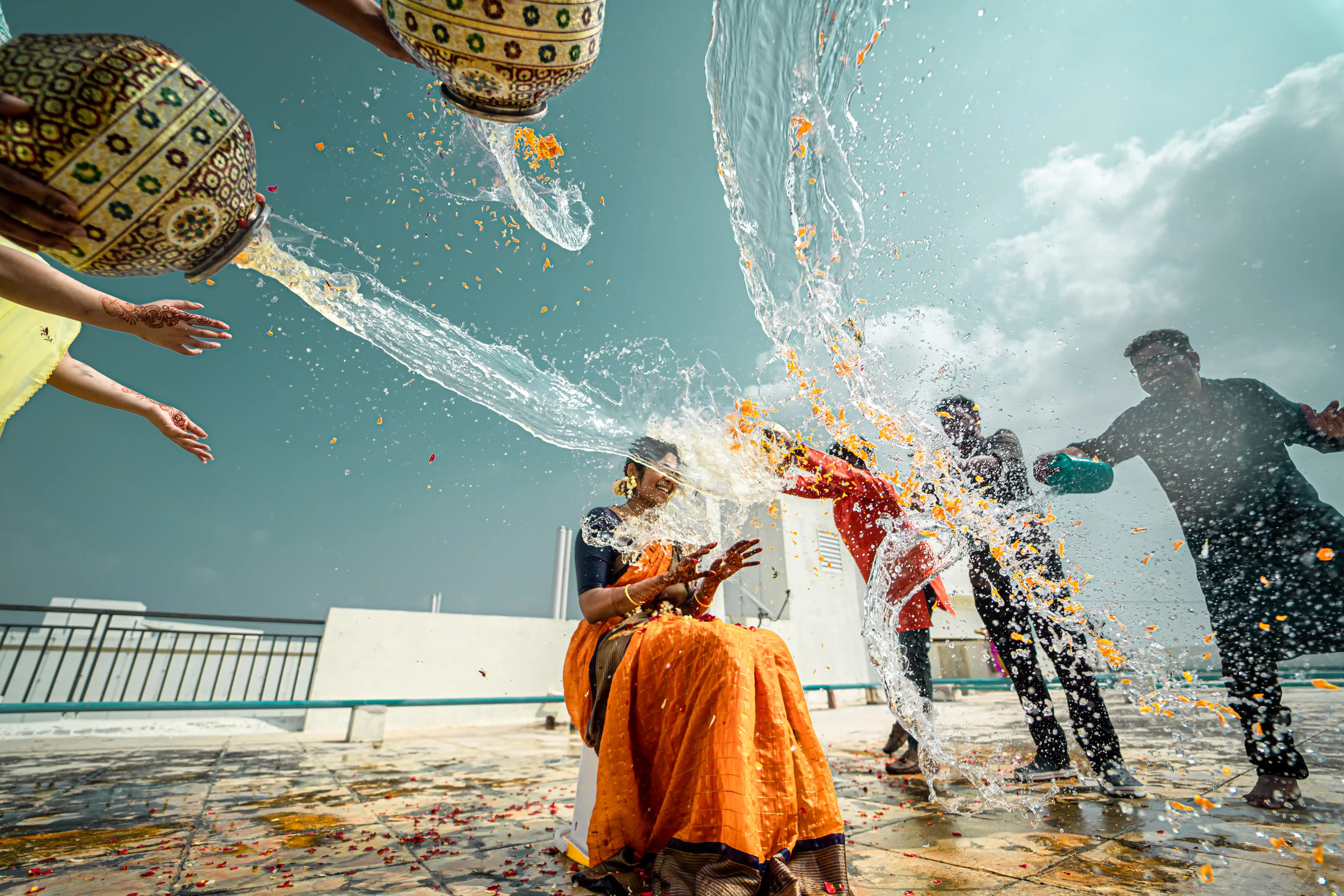 Harshitha in an orange dress, playfully splashed with water during her ‘pellikuturu’ ceremony. Captured by Out of The Blues Fine Art Wedding Photography in Hyderabad.