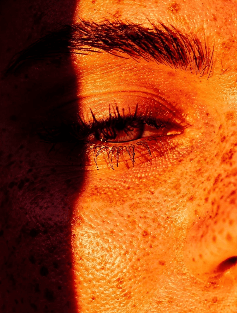 Close-up of a face, emphasizing one eye and brow in warm sunlight. Freckles are visible, with stark shadows creating a dramatic, contemplative mood.