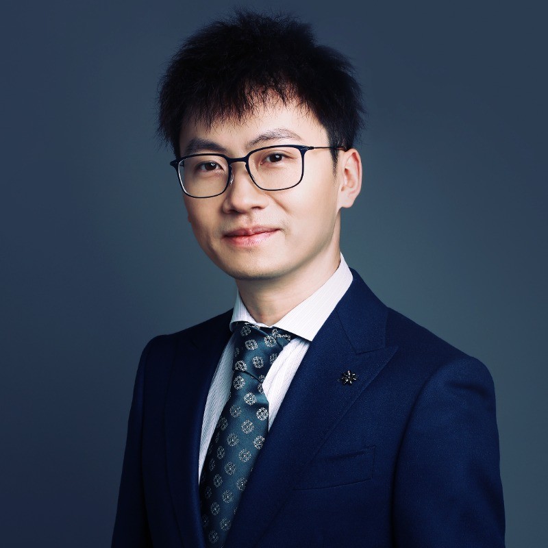 A young man wearing glasses and a formal suit, standing against a plain dark background.