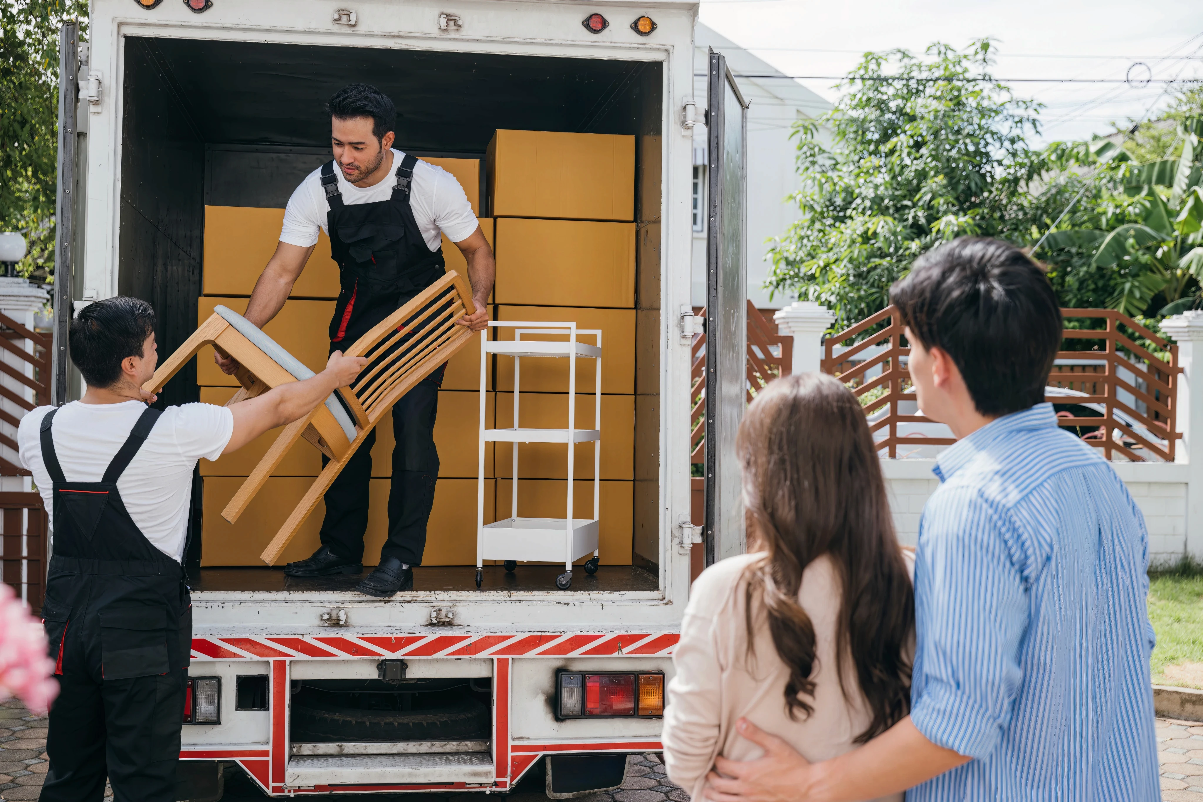 Professional movers carrying a large cardboard box into a house while a couple watches.