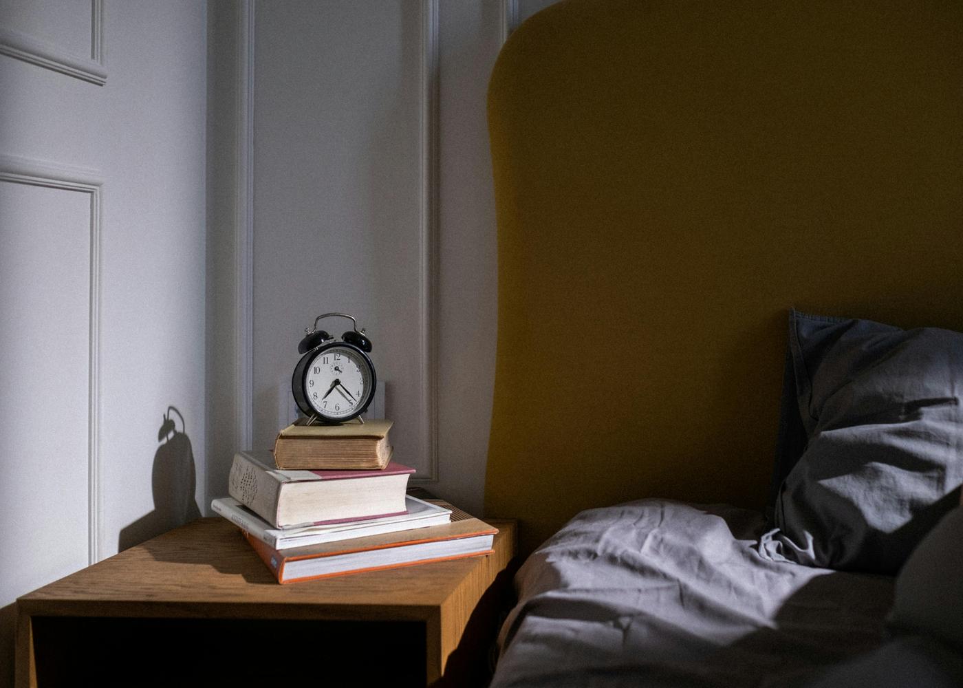 Books stacked on a side table with an alarm clock on top next to a bed