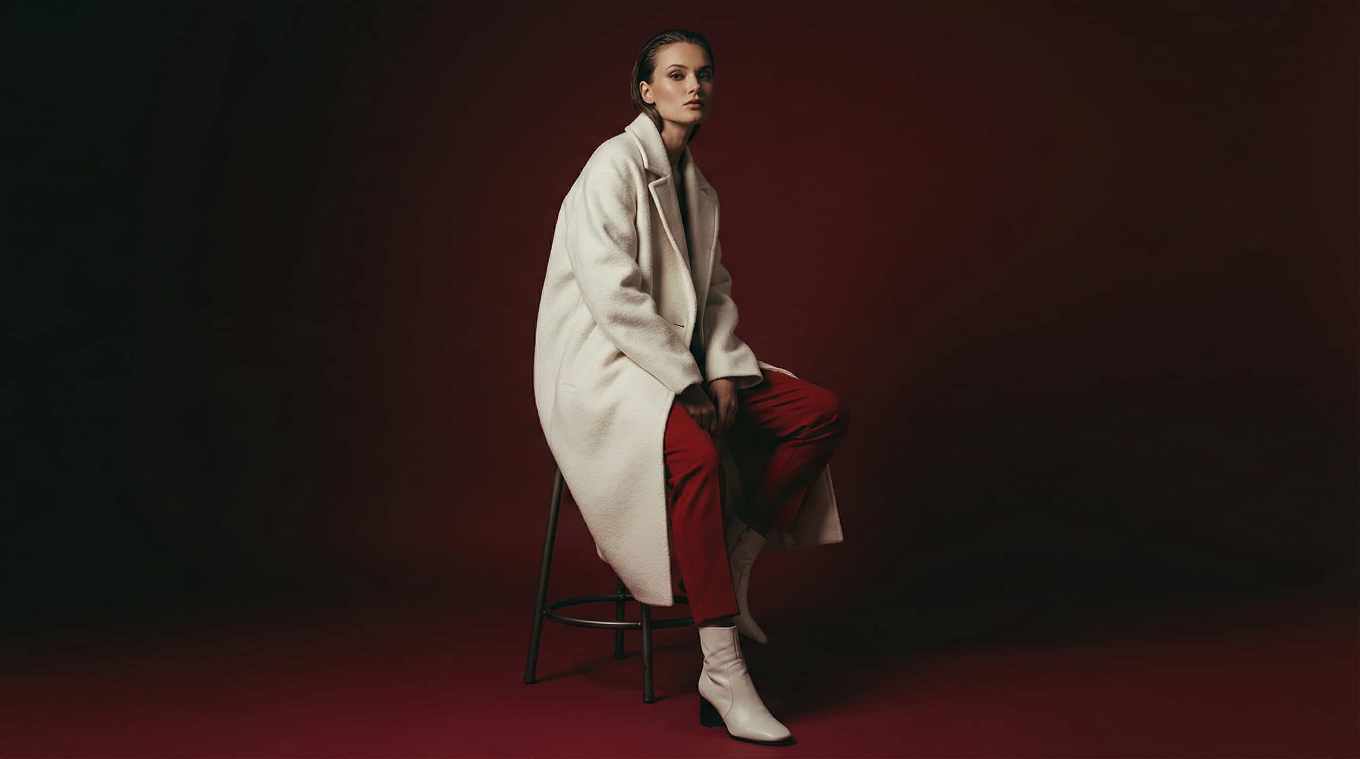 Fashion model seated in a white coat and red outfit on a red background.