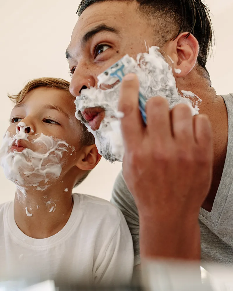 A father and young son with shaving foam on their faces, sharing a playful moment in the bathroom mirror