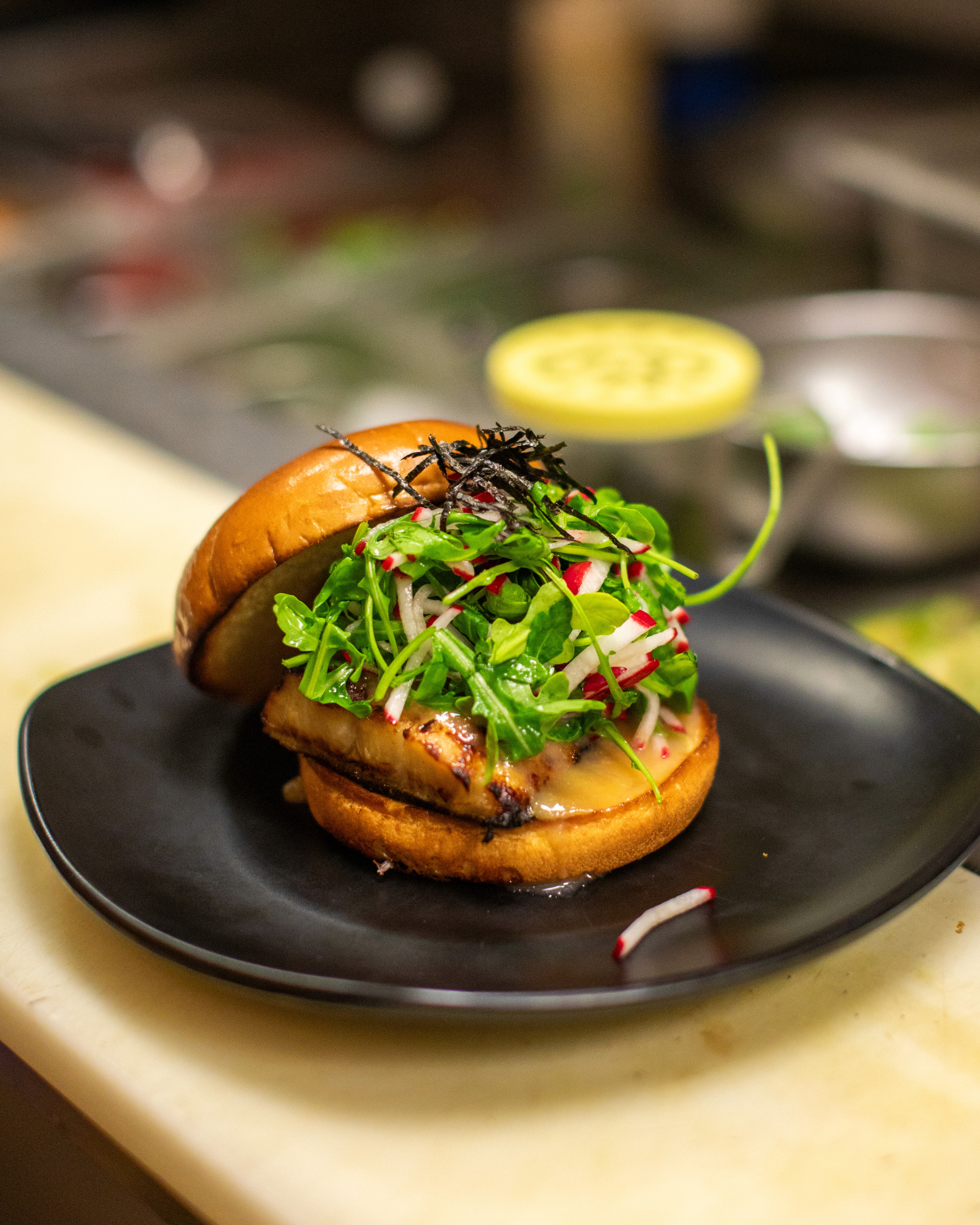 A close-up of a dark gray bowl filled with white fish chunks, cherry tomatoes, green olives, and herbs, served next to a glass of white wine on a white table by a window.