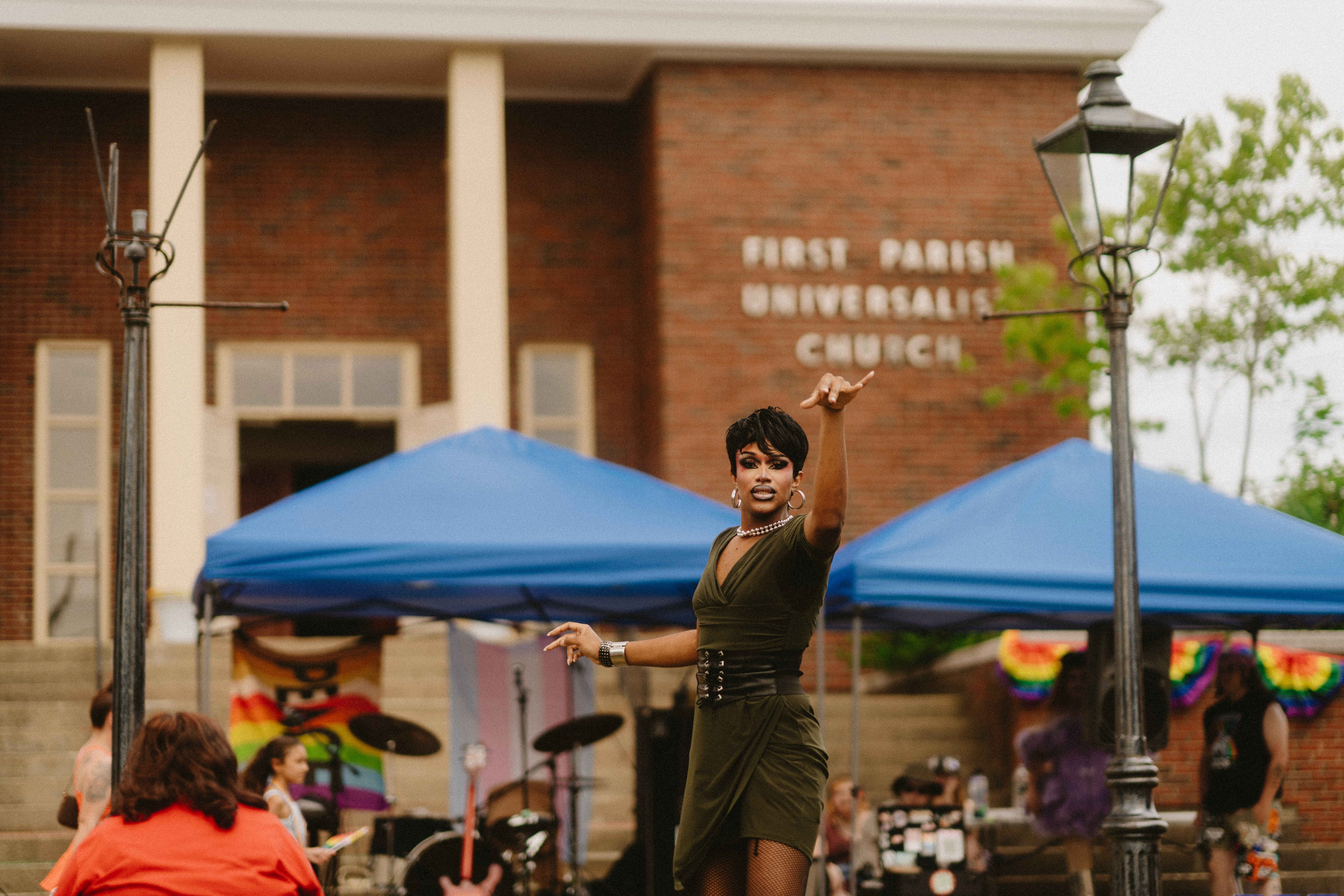 Drag performer Nerukessa mid-performance on the walkway at Stoughton Pride, the First Parish Universalist Church sign clearly visible behind.