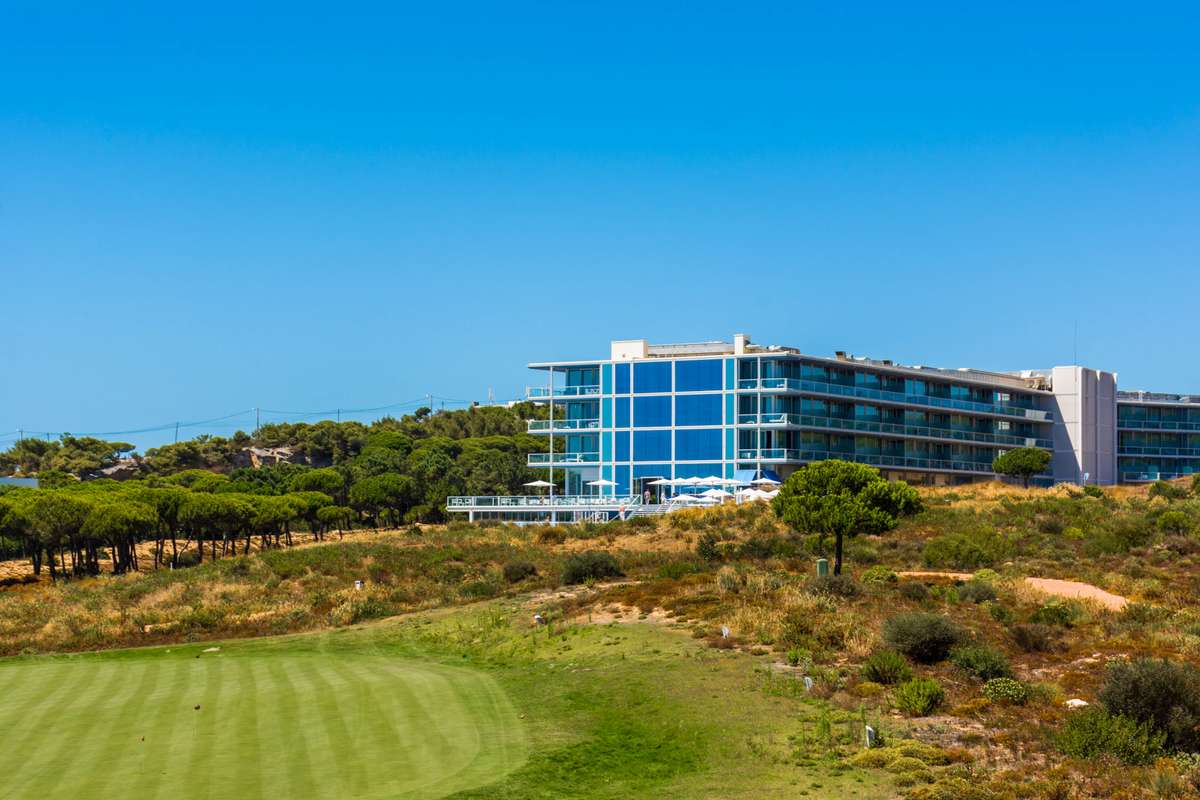 A modern glass-fronted hotel sits behind a green golf course and a line of trees under a clear blue sky.