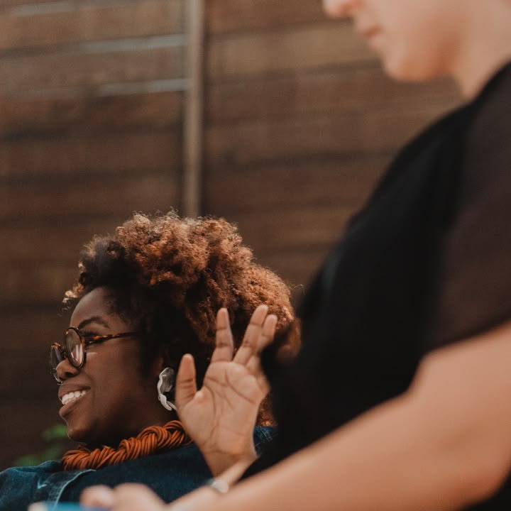 Two people engaging in conversation; one smiles while gesturing with their hand. The setting is casual, with a warm, relaxed ambiance.