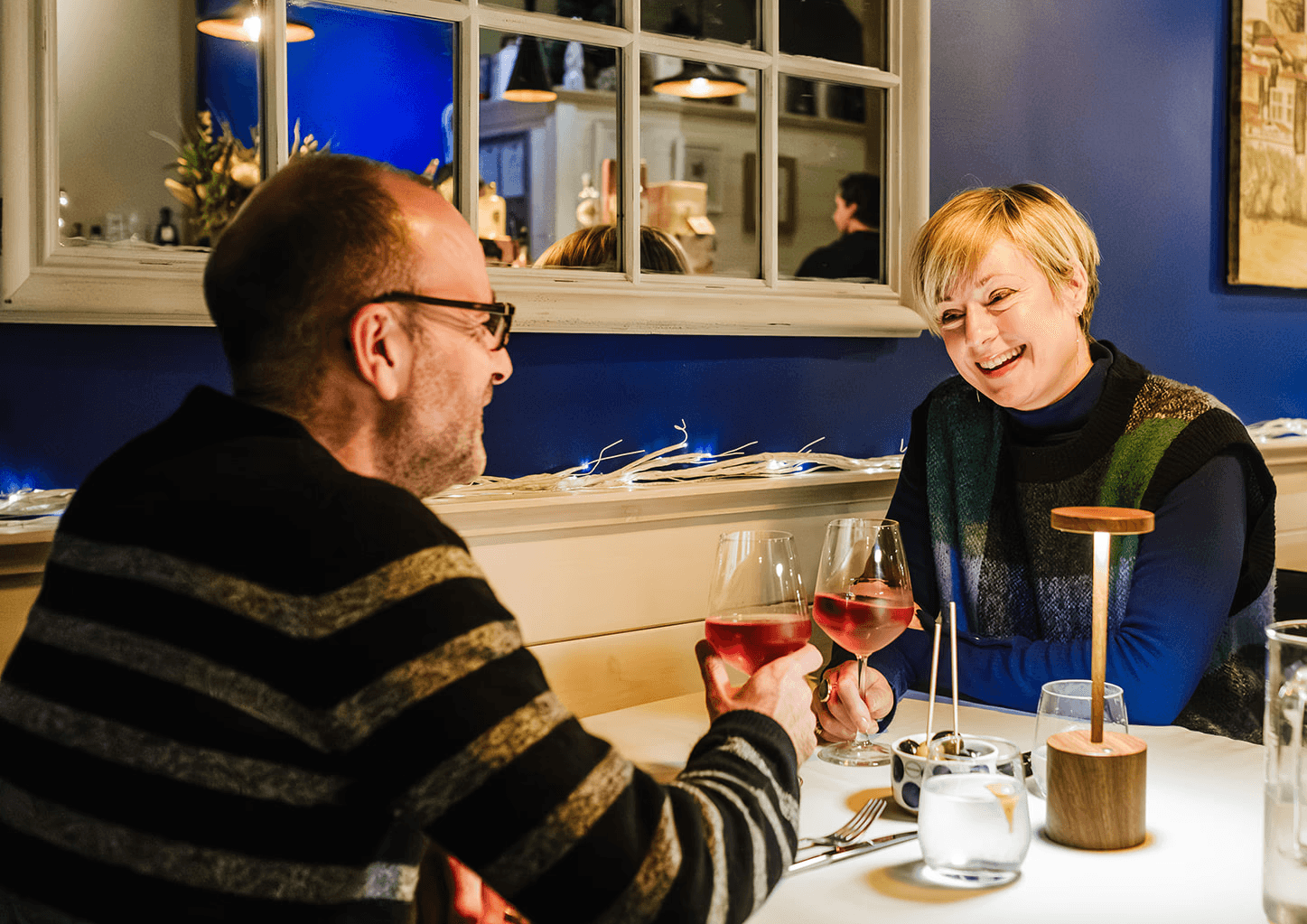 A man and woman enjoying a meal together in a warmly lit restaurant, smiling as they clink glasses of rosé wine. Decorative lights run along the wall behind them, and a small lamp and bowl of olives sit on their table.