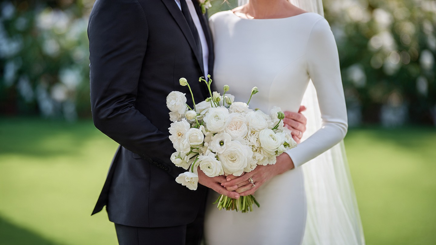 Elegant bridal couple captured from the waist up in a garden setting; the groom in a dark tailored suit with boutonniere embraces the bride in an off-shoulder white mermaid gown, both holding a large cascading bouquet of creamy white ranunculus and greenery; soft natural light filters through.