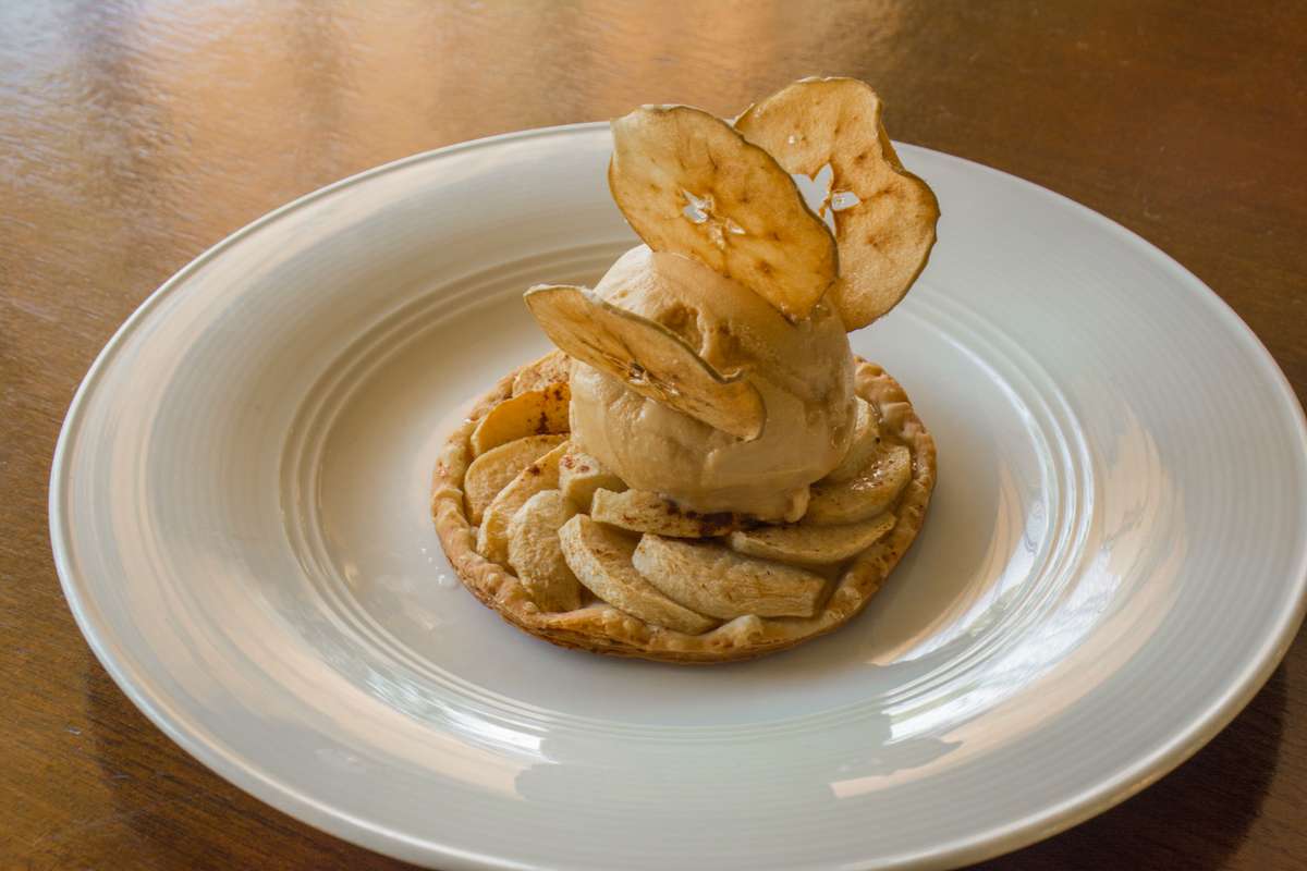 A seared scallop topped with a thin slice of roasted meat and herbs sits on a round cracker atop a white plate.