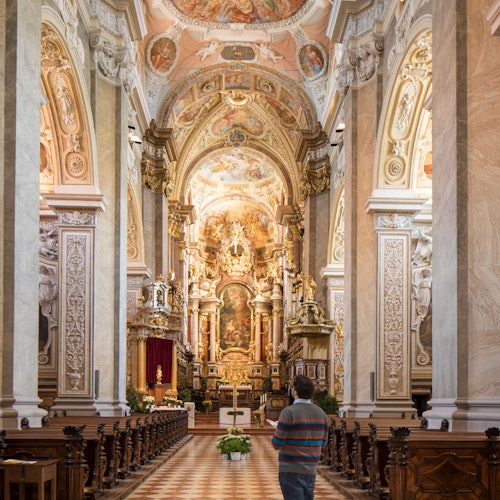 An ornate baroque church interior with intricate carvings and paintings, featuring pews and an altar with a person admiring the view.