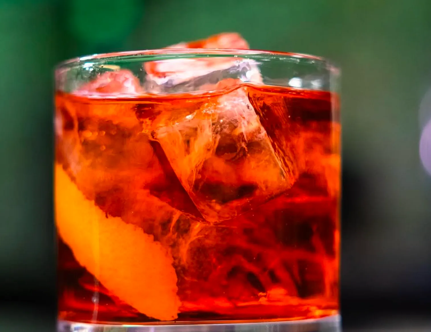 Close-up of a bright amber-red cocktail in a short glass filled with ice, featuring a large orange peel garnish against a softly blurred green and dark background.