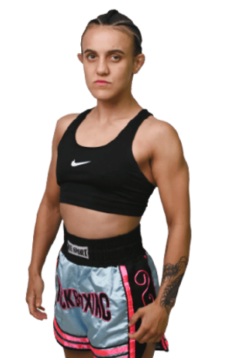 A female kickboxer in a black sports bra and silver shorts with pink accents stands confidently against a plain background, showcasing her athletic physique and braided hairstyle.