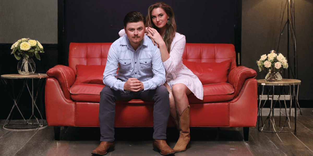 Colin and Kathleen Ray, a bluegrass duo, seated on a red couch with a serene backdrop, showcasing their musical partnership.