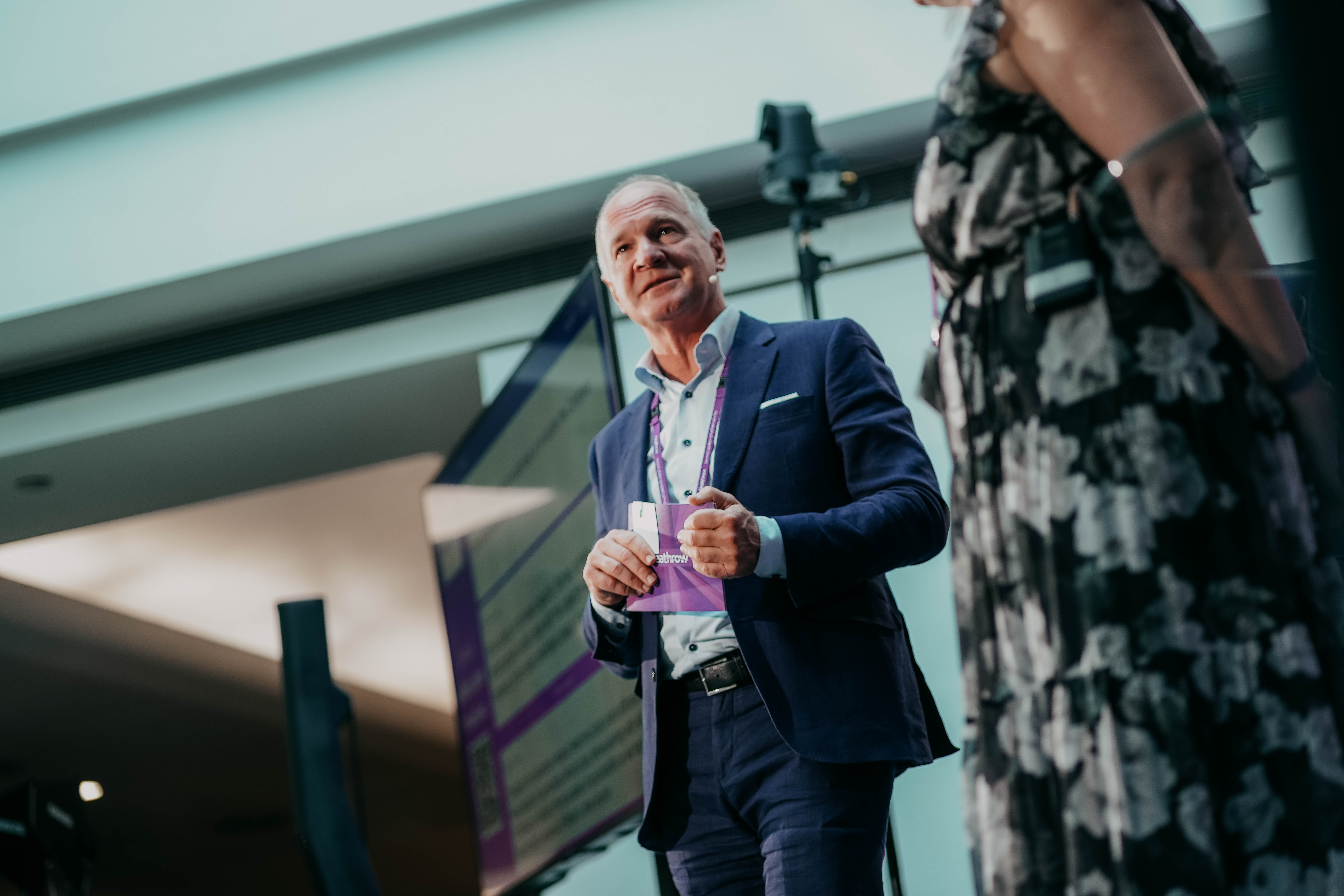 A low-angle shot of Heathrow CEO Thomas Woldbye speaking on a stage during a presentation. He is wearing a blue suit, holding a pamphlet, and looking towards someone just out of frame.