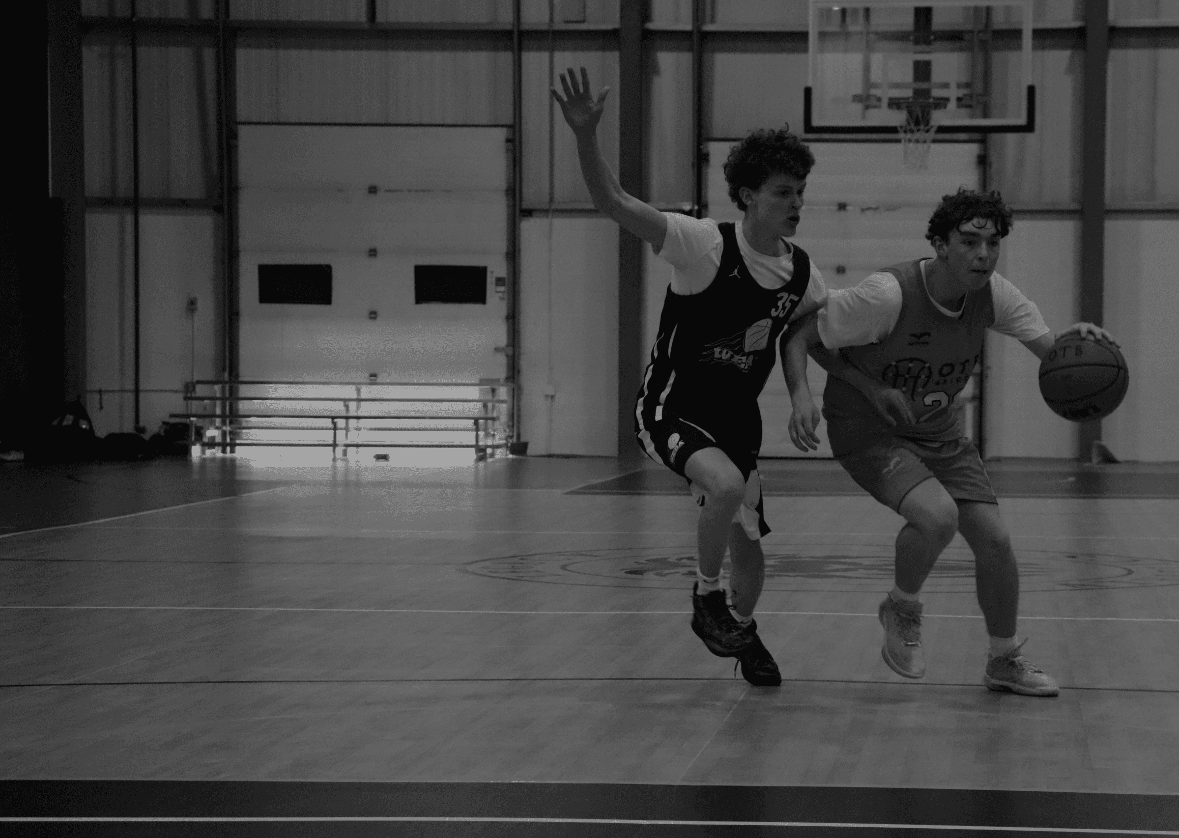 A youth basketball player in an orange jersey dribbling aggressively past a defender during a game, with a referee and spectators visible in the background on an indoor court.