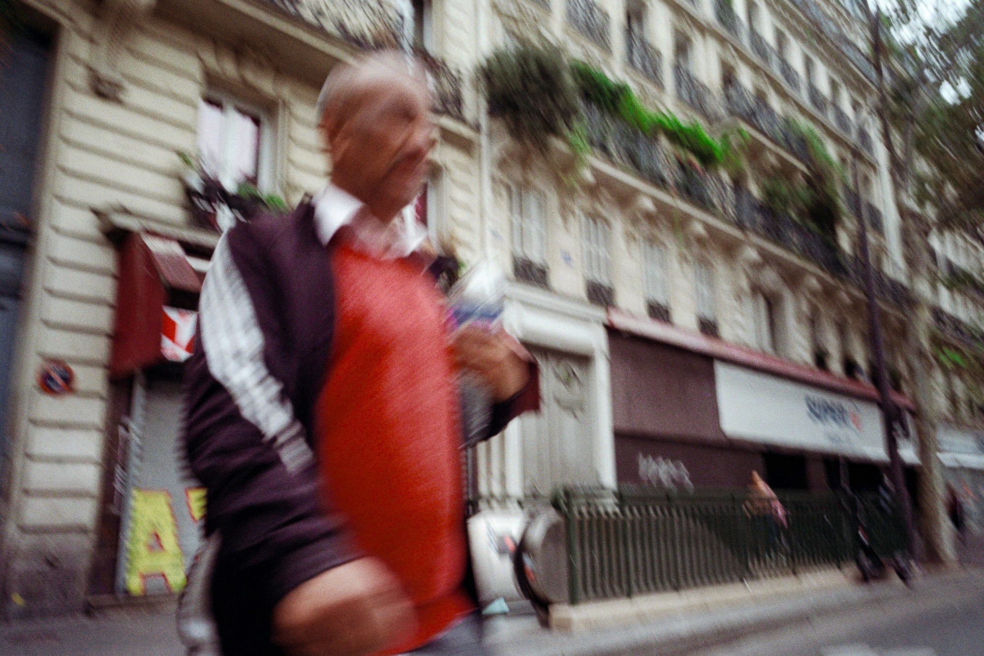A man with a blurred appearance walks past a row of classic Parisian buildings featuring ornate balconies and greenery, holding a water bottle, with a bicycle visible in the urban background.