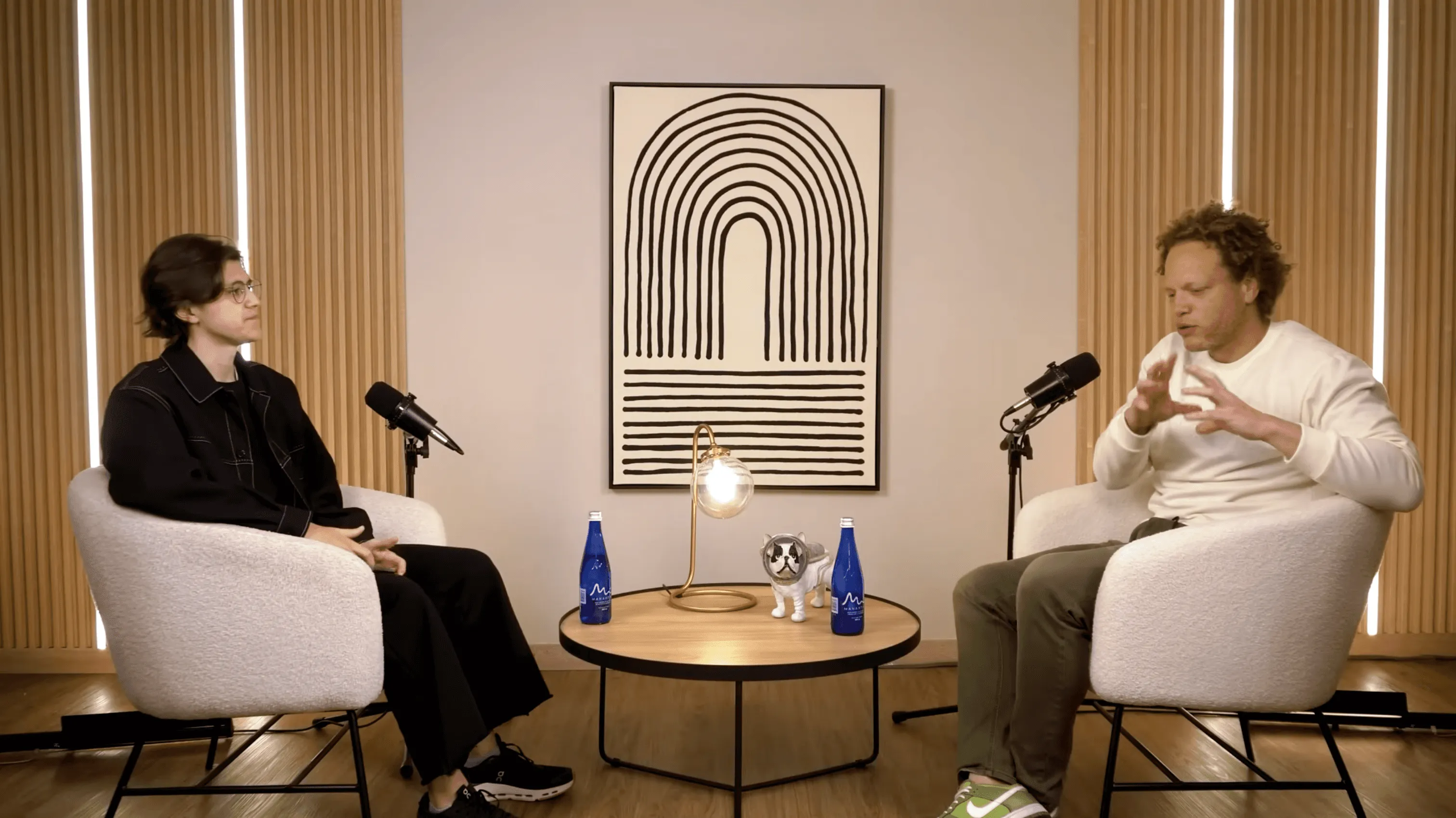 Dos personas sentadas frente a frente en un estudio de grabación, conversando con micrófonos sobre una mesa central, en un espacio minimalista con iluminación cálida y arte abstracto al fondo.