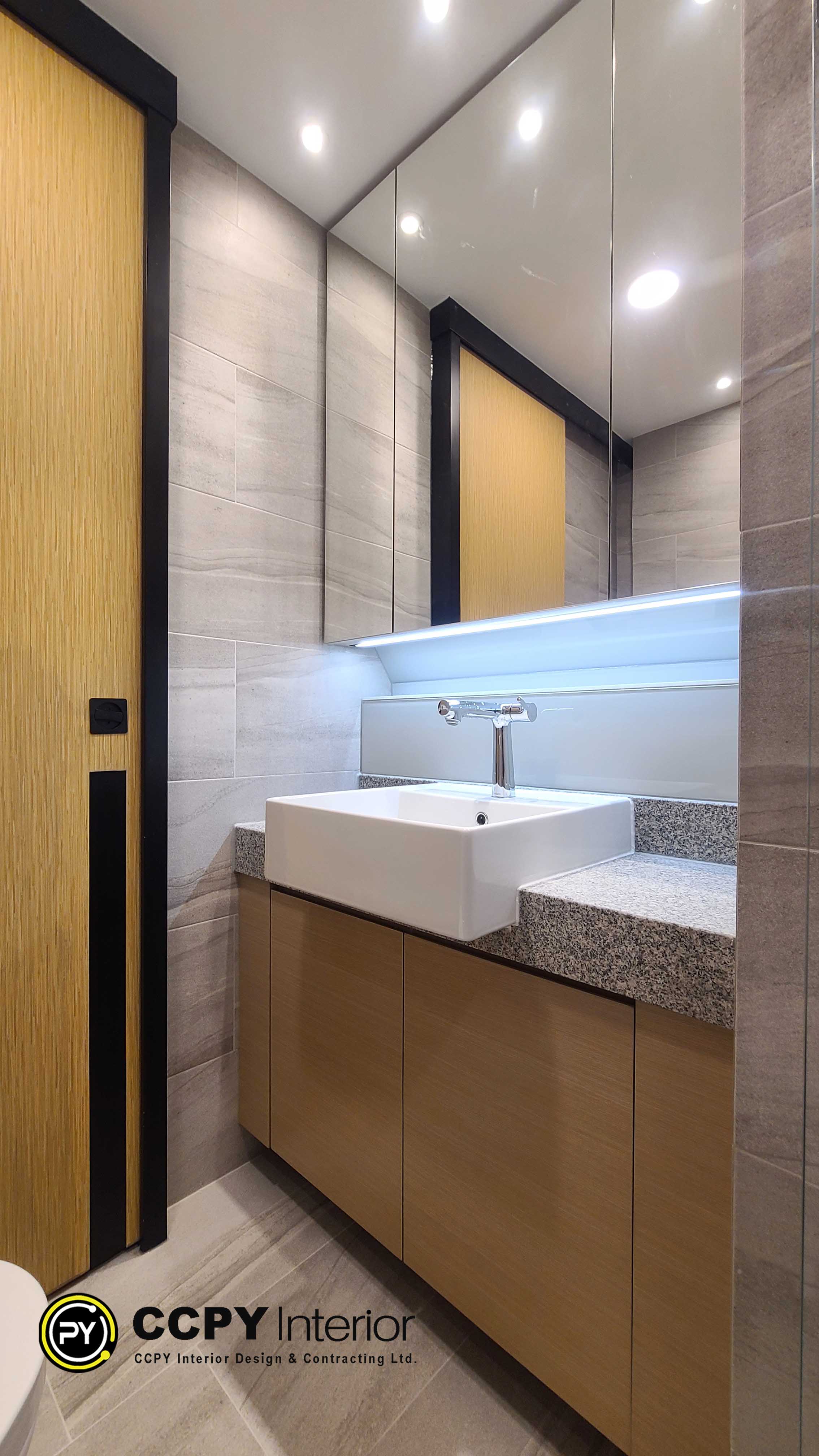 This is a modern bathroom vanity featuring a white rectangular vessel sink and an illuminated mirror cabinet.The vanity area is designed with light-colored, textured stone tiles and sleek, minimalist fixtures.Integrated LED lighting is embedded beneath the mirror cabinet for functional and ambient lighting.The cabinet features a sliding door with a light wood grain finish, commonly used in contemporary Hong Kong residential interior designs.