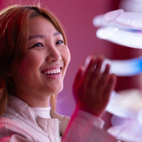 A person smiles with their hand raised near a colorful, illuminated spiral structure on a pink and red background.