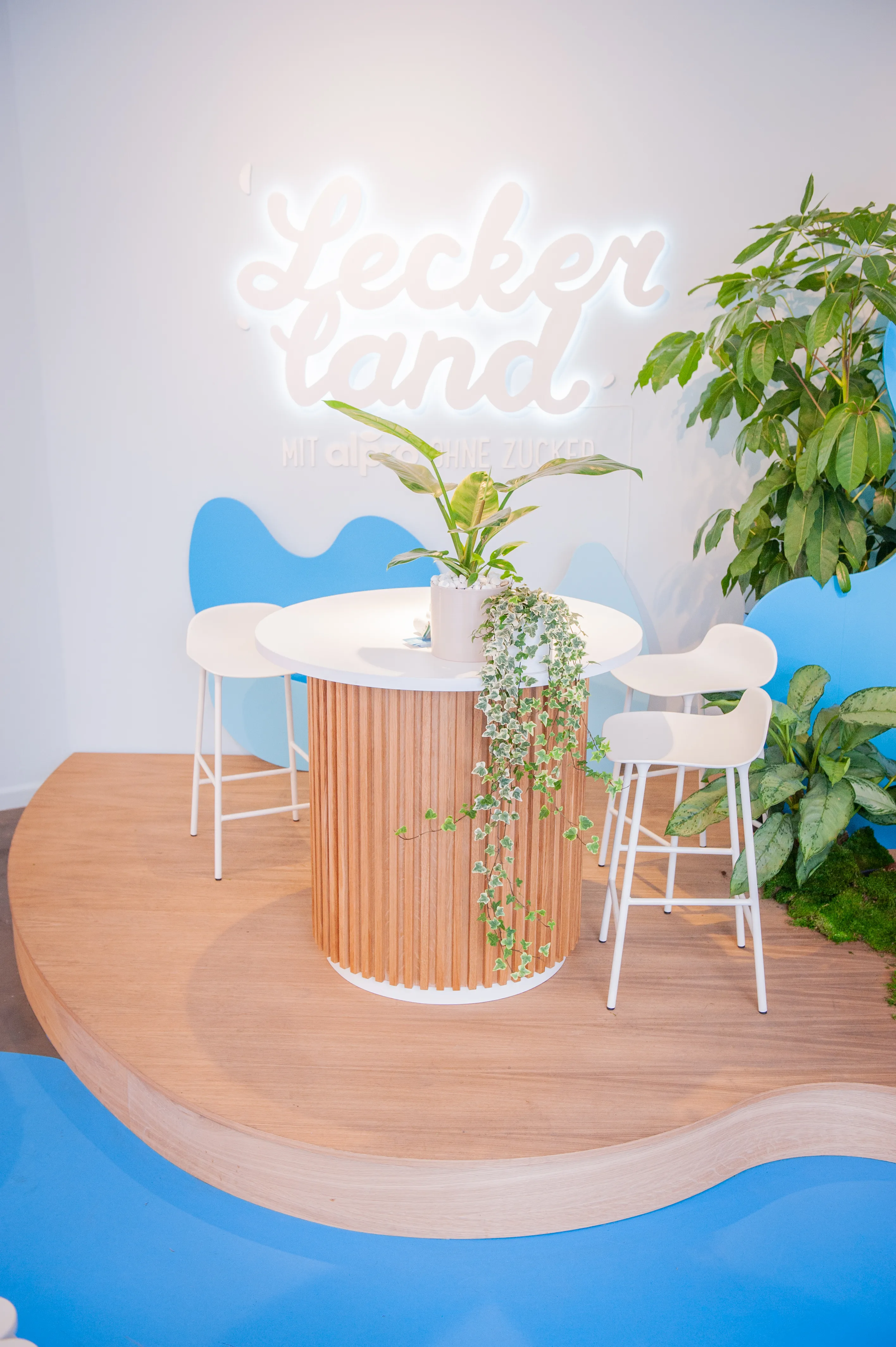 High table with wood-slat base and white bar chairs beneath the glowing Leckerland neon logo at the Alpro pop-up.